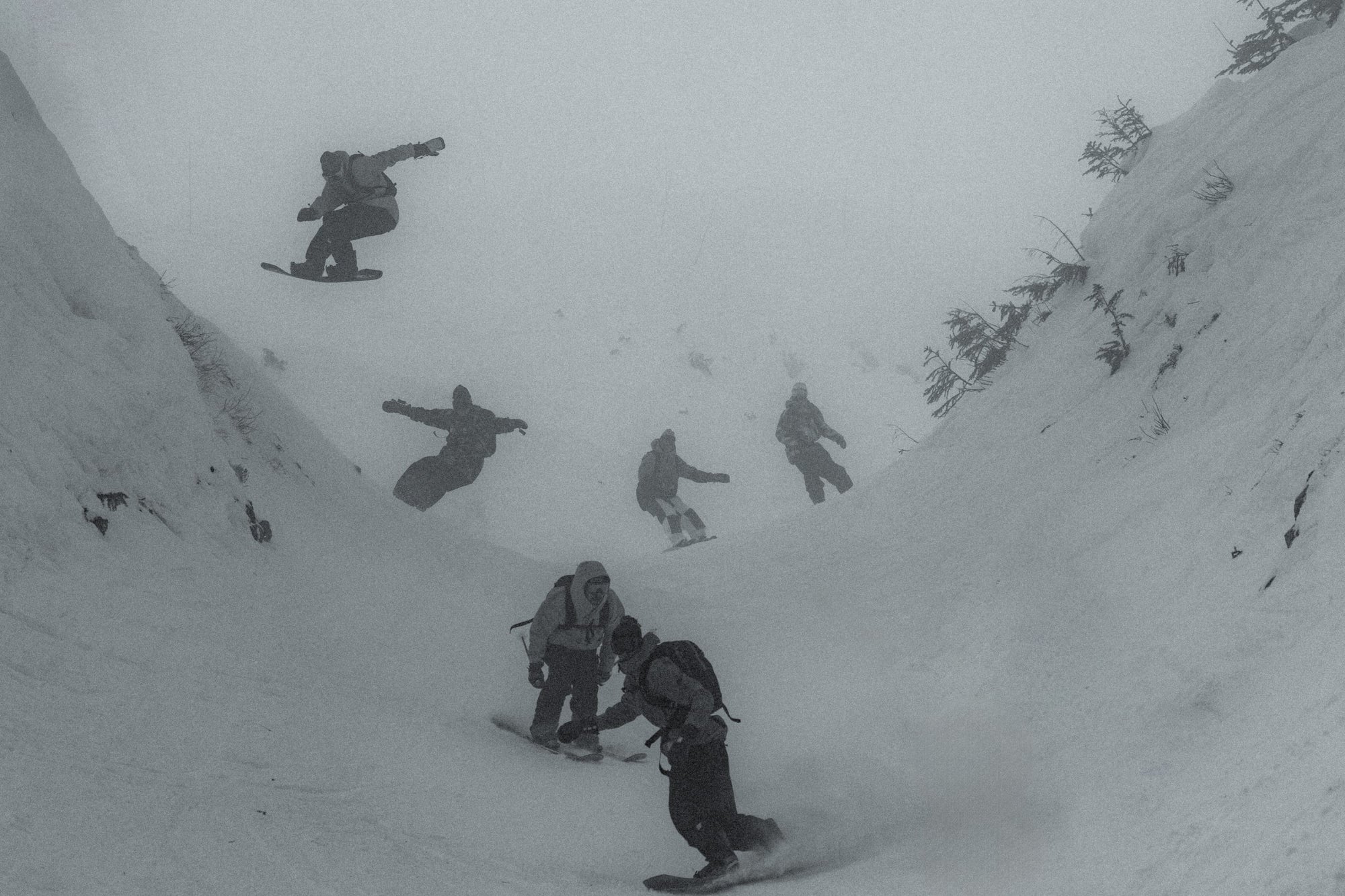 A group of snowboarders and skiers riding down a U-shaped valley on a cloudy, snowy day.