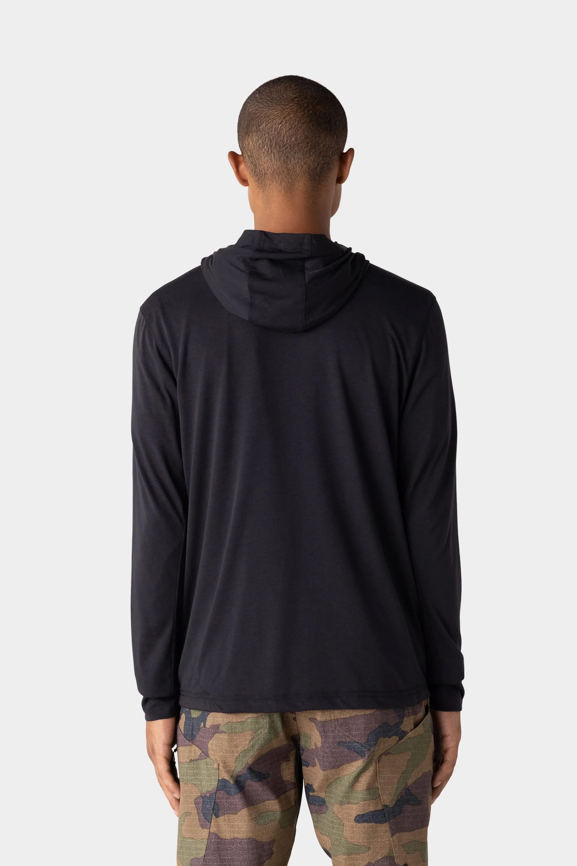 A person is shown from the back wearing a 686 Mens Everywhere drirelease® Long Sleeve Hooded Tech Tee in black with camouflage pants, standing against a plain light gray background.