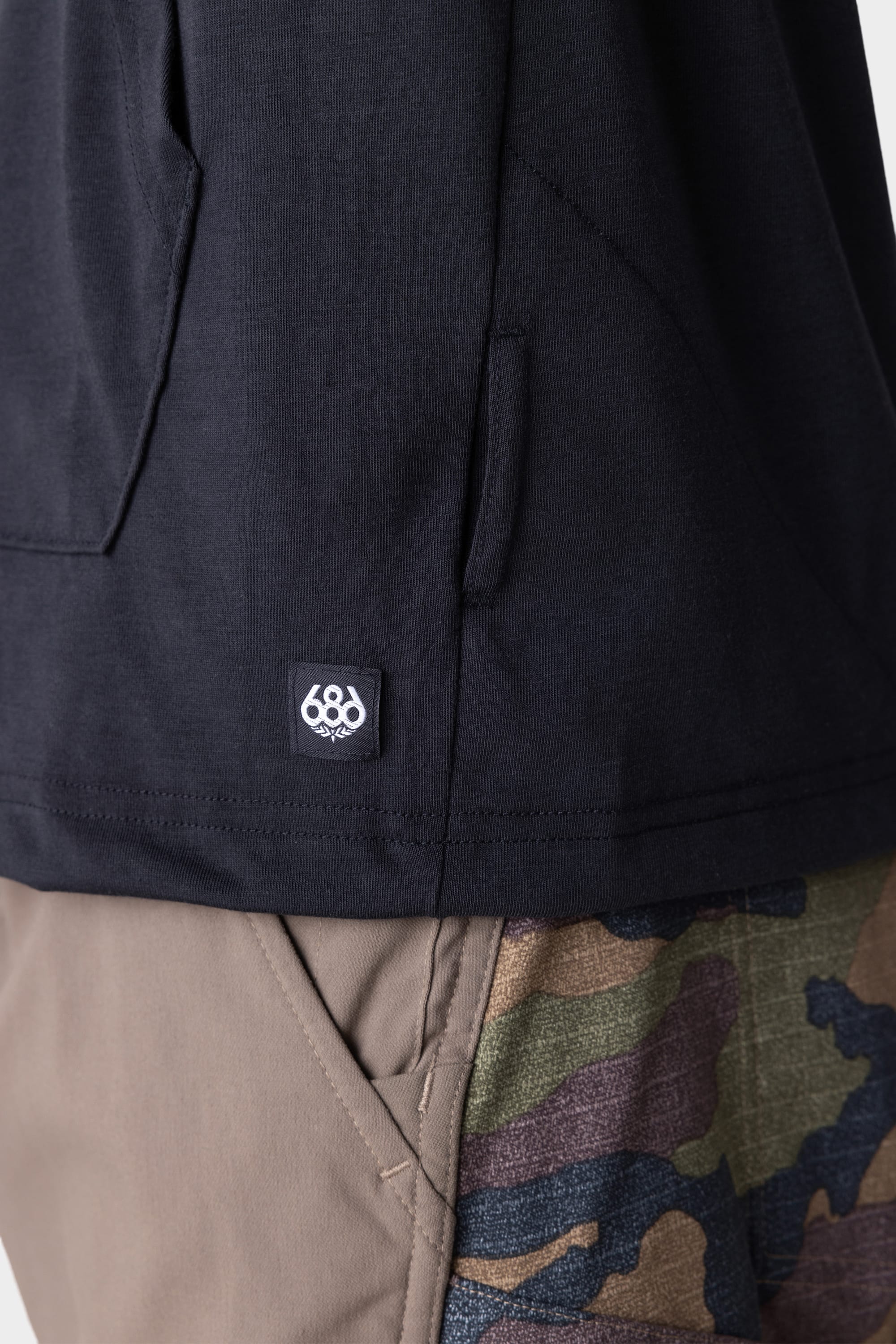 Close-up of a person wearing the 686 Mens Everywhere drirelease® Long Sleeve Hooded Tech Tee in black, featuring a small 686 logo tag near the hem, paired with pants that have solid tan and camouflage panels.
