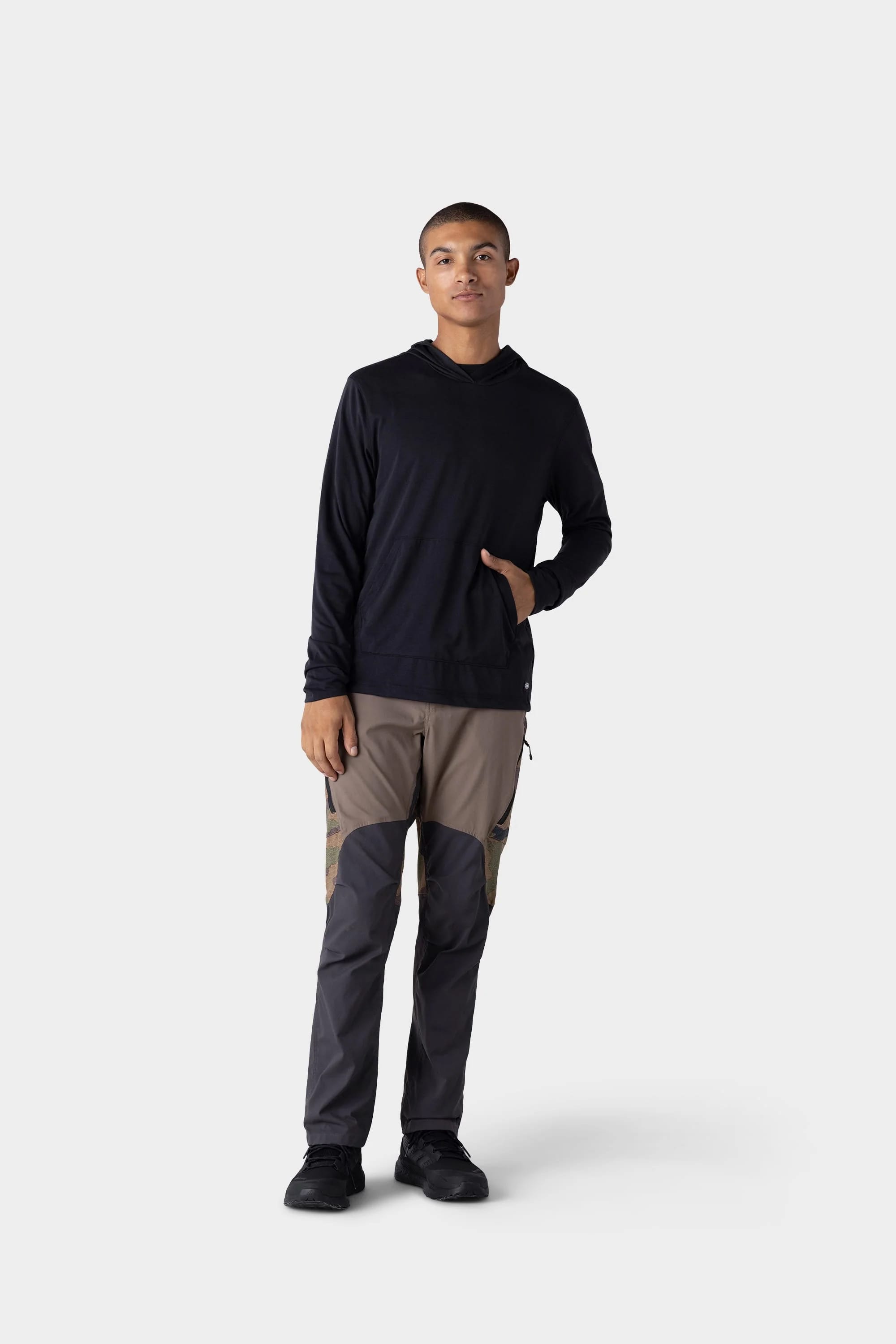 A person stands against a light gray background, wearing the 686 Mens Everywhere drirelease® Long Sleeve Hooded Tech Tee in black, with brown and gray color-blocked pants and black shoes, one hand in the hoodie’s front pocket.