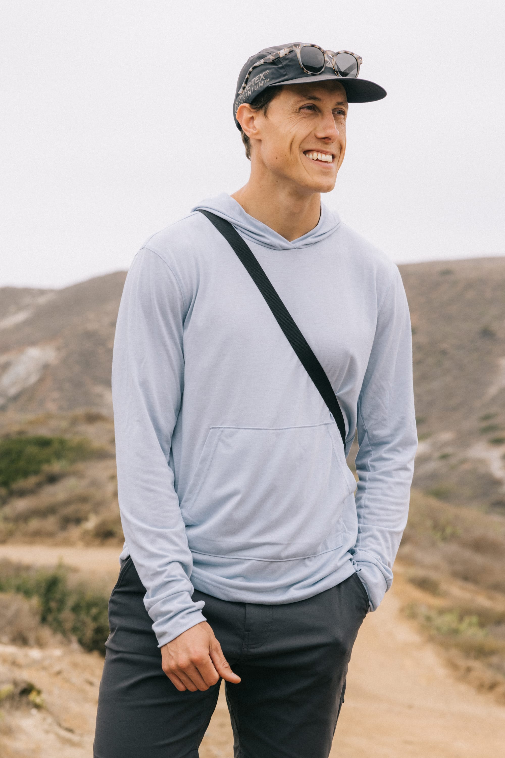 A smiling man wears the 686 Mens Everywhere drirelease® Long Sleeve Hooded Tech Tee, black pants, and a cap with sunglasses on top while standing on a dirt trail in a hilly outdoor landscape.