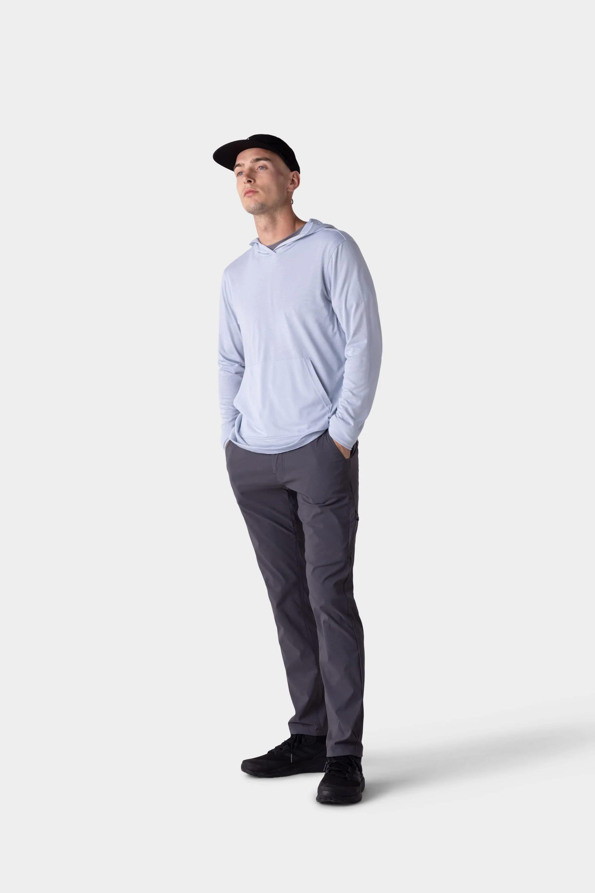 A man stands against a plain white background, wearing the 686 Mens Everywhere drirelease® Long Sleeve Hooded Tech Tee by 686, paired with dark gray pants, black sneakers, and a black cap. His hands are in his pockets as he gazes slightly upward.