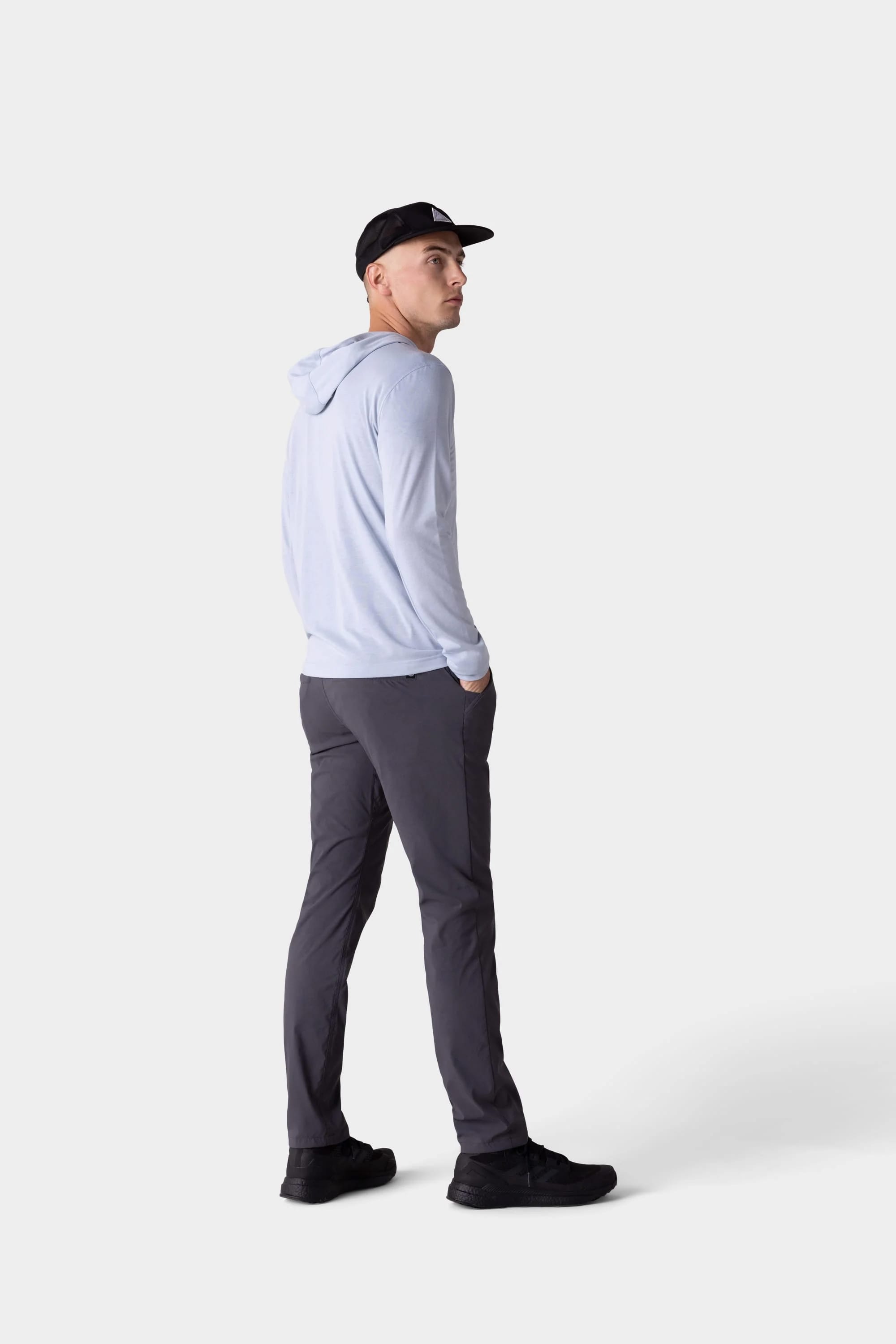A man wears the 686 Mens Everywhere drirelease® Long Sleeve Hooded Tech Tee by 686 with dark gray pants, black shoes, and a black cap, standing with hands in pockets against a plain light background.