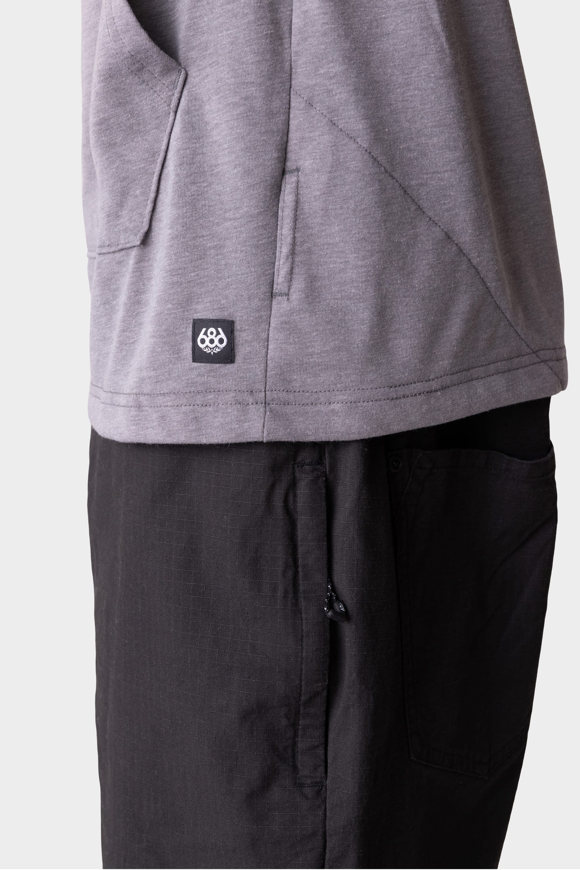A close-up side view shows a person wearing the 686 Mens Everywhere drirelease® Long Sleeve Hooded Tech Tee in gray, featuring a small 686 logo tag above black pants, highlighting pocket details on both garments against a plain background.