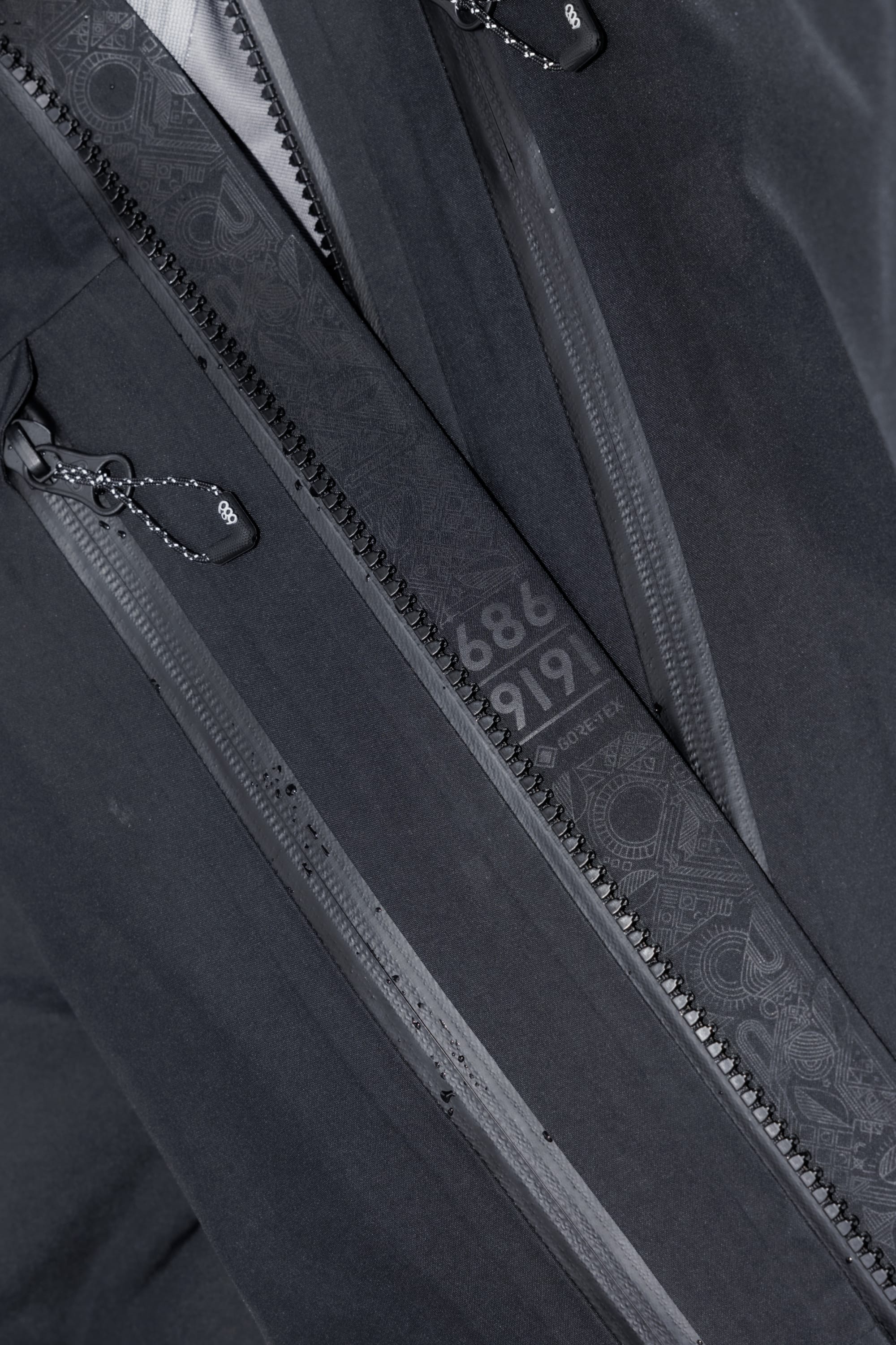 Close-up of the 686 Mens GORE-TEX 3L ATV Jacket in black, crafted from recycled fabric. Features dual zippers with decorative pull cords and an inner flap with subtle patterns plus light grey 686 and 9191 detailing.