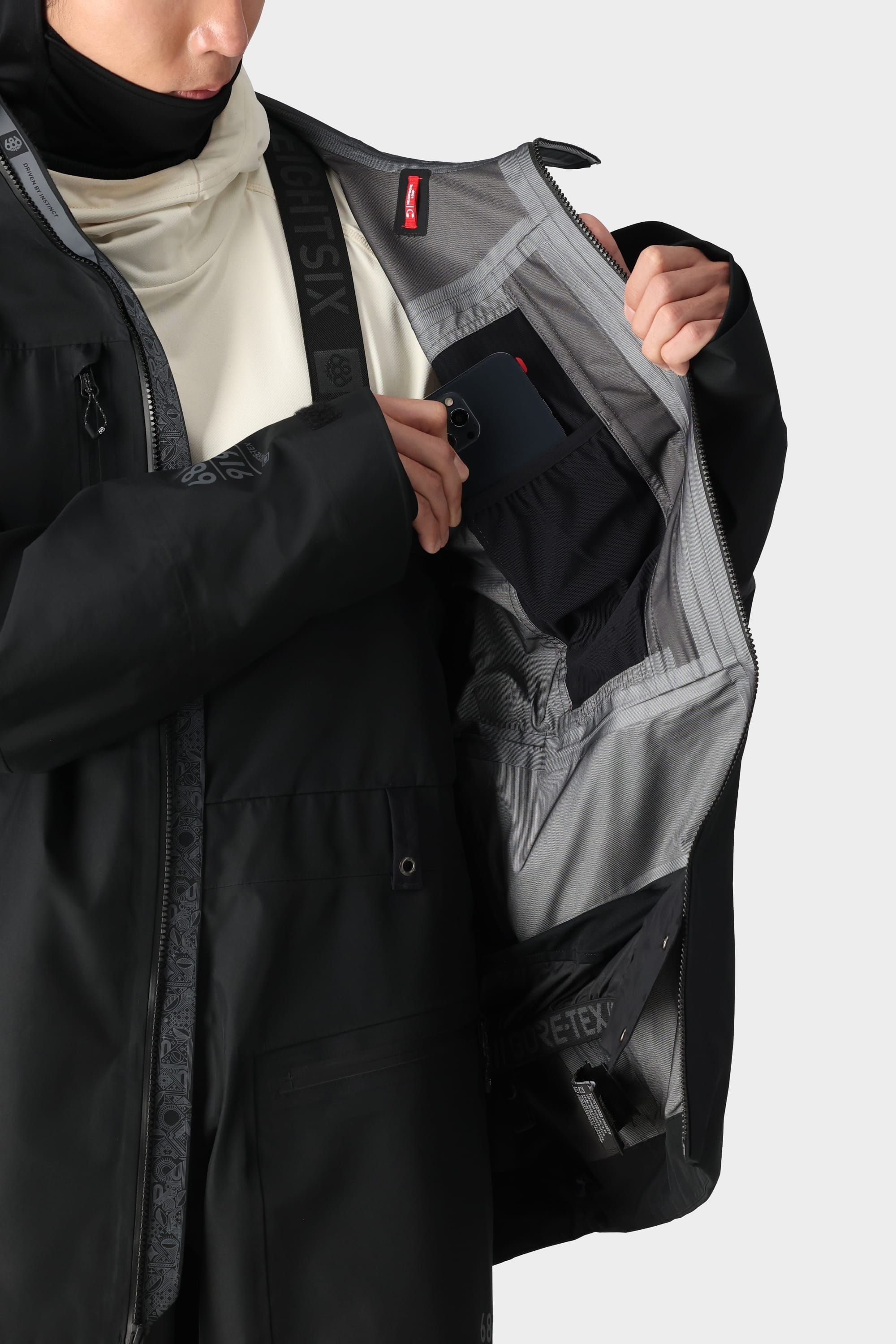 A man in the 686 Mens GORE-TEX 3L ATV Jacket opens the inner pocket, revealing organized compartments and stashes his phone. The light grey recycled lining features multiple pockets for easy organization.