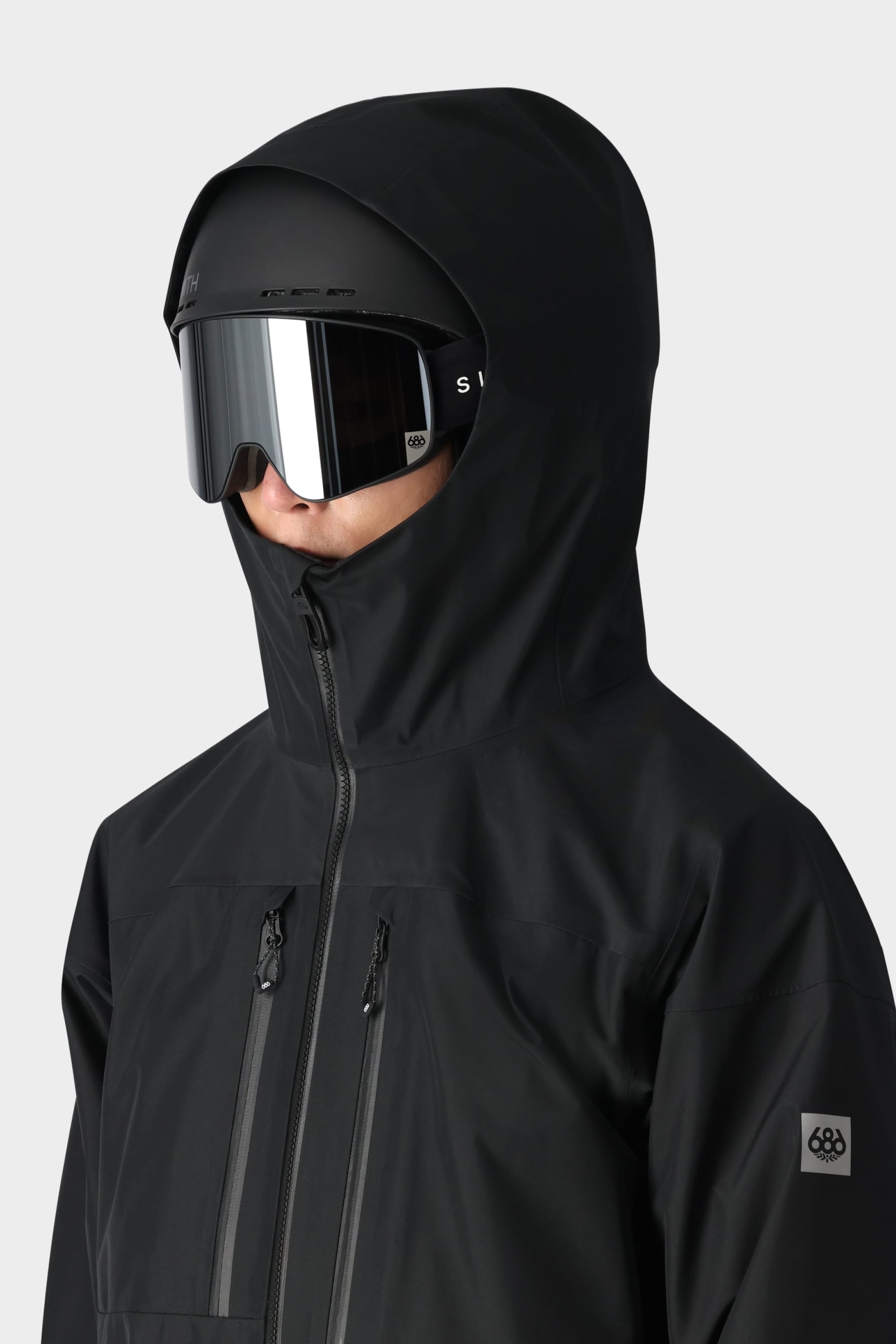 A person wears the 686 Mens GORE-TEX 3L ATV Jacket with the hood up, black ski goggles, and a black helmet, facing slightly right against a plain light background—ideal gear for all-terrain vehicle riders.