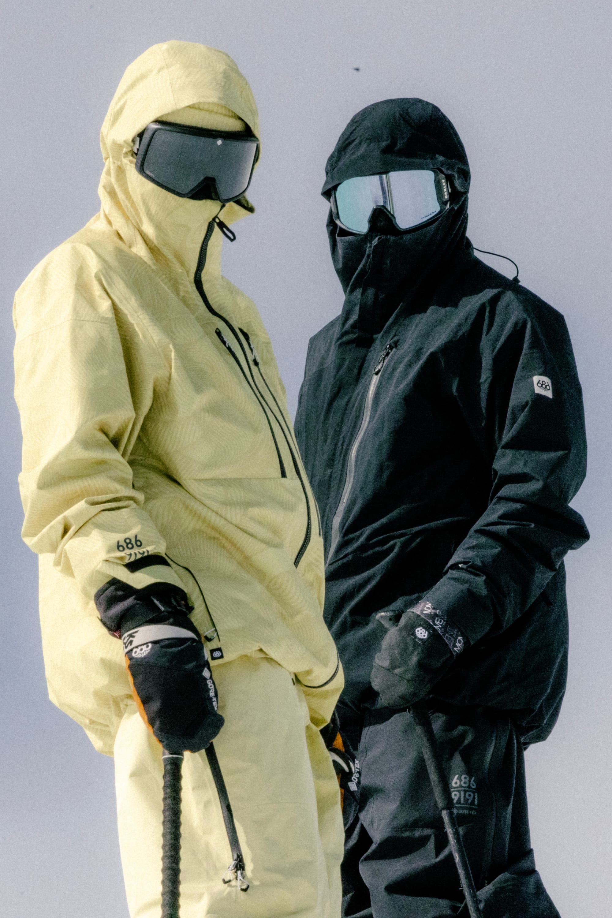 Two people stand side by side against a plain light background, both in winter sports gear and ski poles—one in yellow, one in black—wearing the 686 Mens GORE-TEX 3L ATV Jacket by 686, plus ski goggles and gloves.