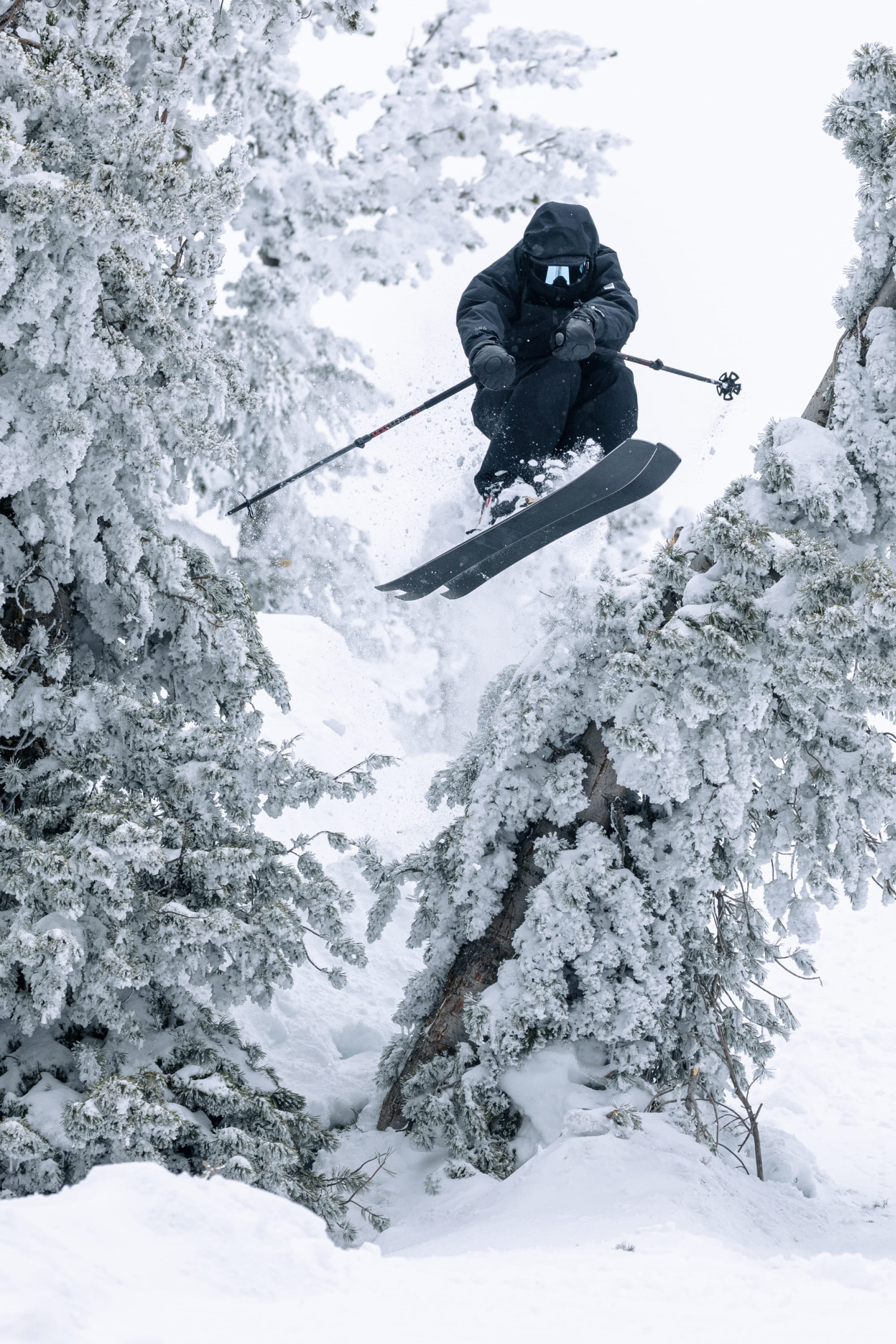 A skier wearing the 686 Mens GORE-TEX 3L ATV Jacket is airborne between snow-covered trees, gripping ski poles and sporting goggles as snow sprays around them.