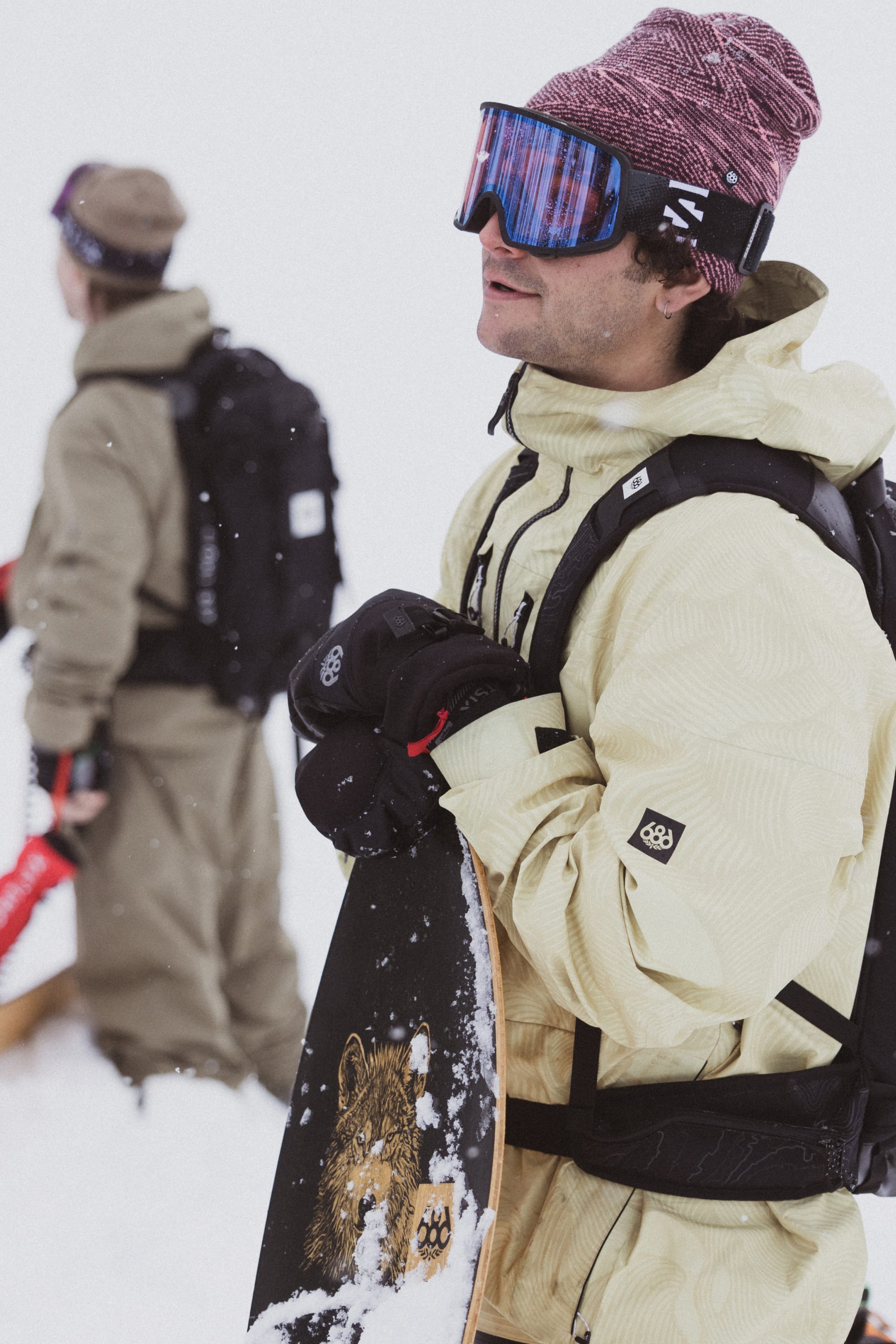 A snowboarder in the 686 Mens GORE-TEX 3L ATV Jacket, gloves, and a purple beanie holds a snowboard with a lion graphic. Another snowboarder stands in the background amid snow.