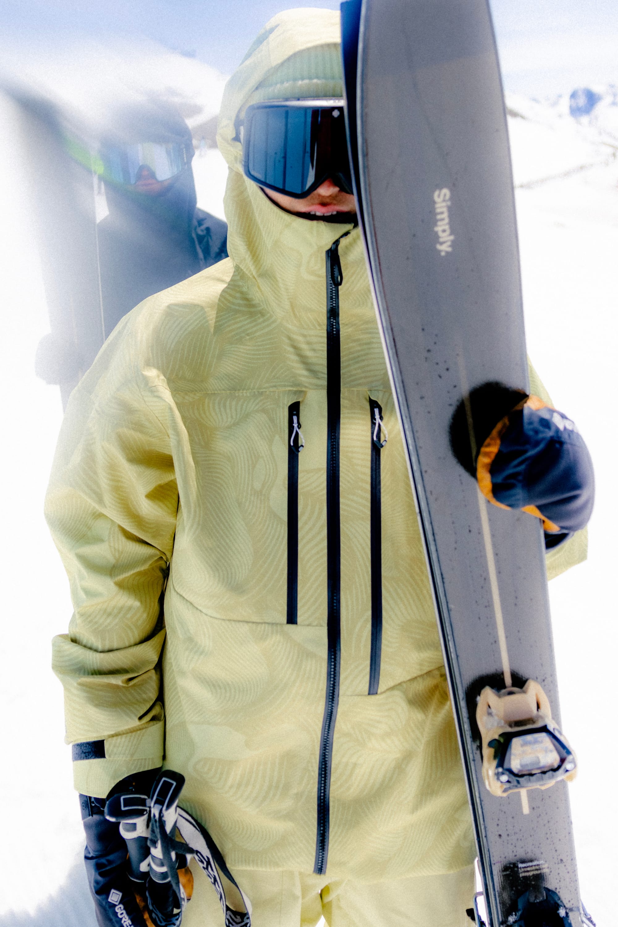 Wearing a yellow 686 Mens GORE-TEX 3L ATV Jacket, black gloves, ski goggles, and a hood, a skier holds skis upright on a snowy mountain while another skier is partially visible in the background.