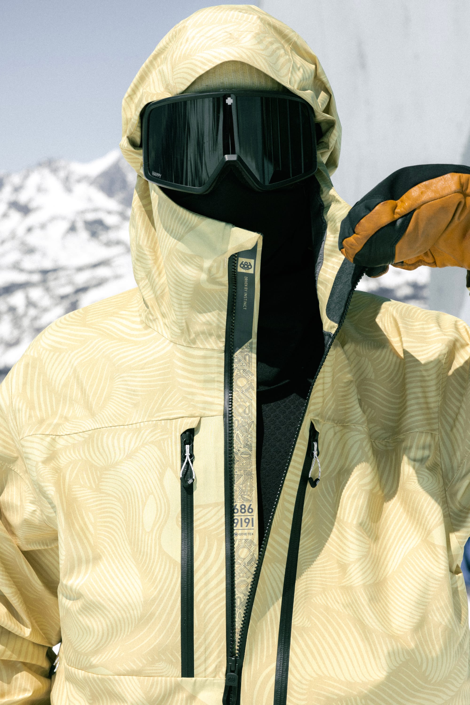 A person wearing the 686 Mens GORE-TEX 3L ATV Jacket, trusted by all-terrain vehicle riders, stands outdoors in a snowy mountain setting zipping up their yellow patterned jacket with black ski mask, large goggles, and brown gloves.