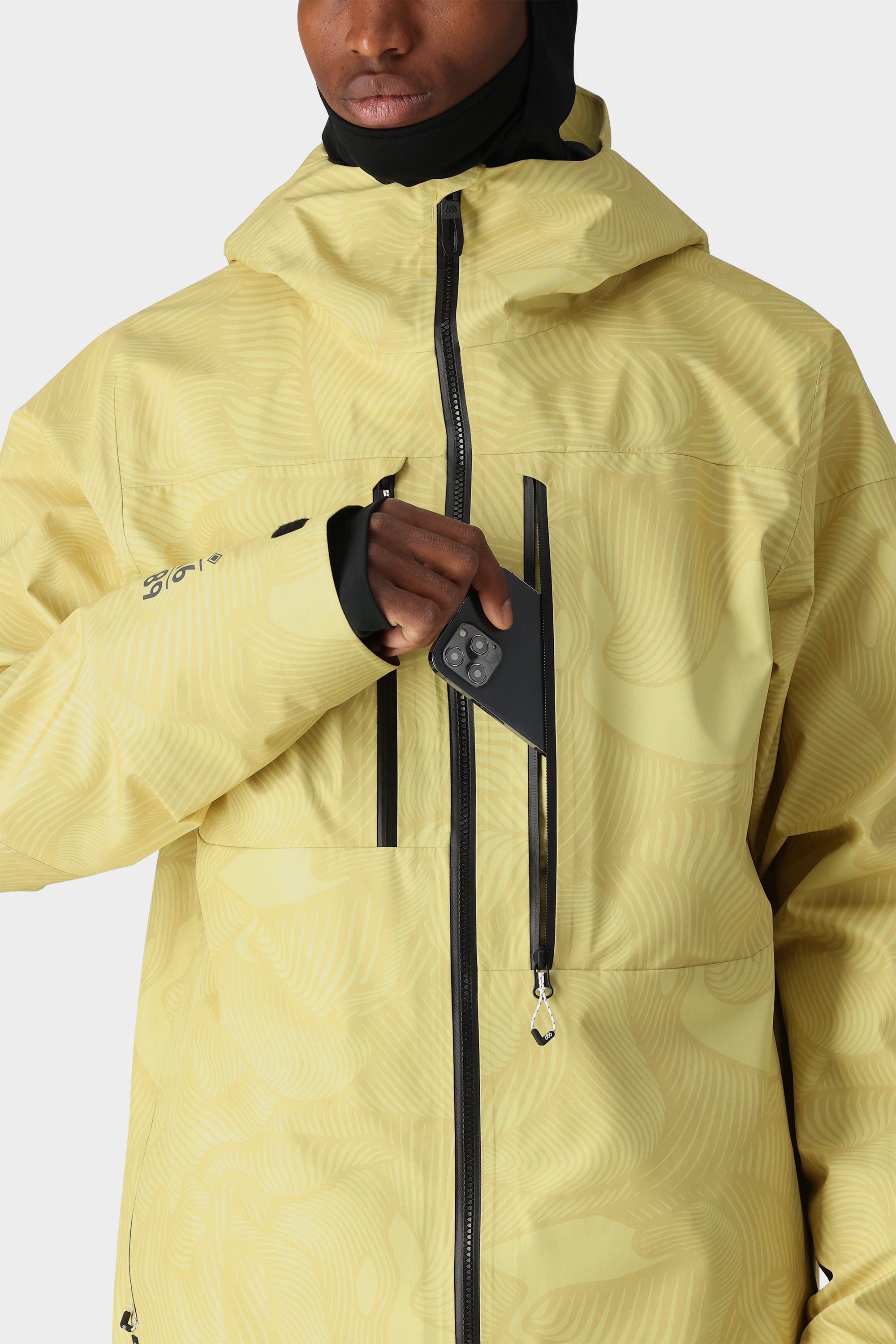 A person, their lower face visible, places a smartphone into the chest pocket with a vertical zipper on the 686 Mens GORE-TEX 3L ATV Jacket by 686, featuring a yellow patterned hooded design.