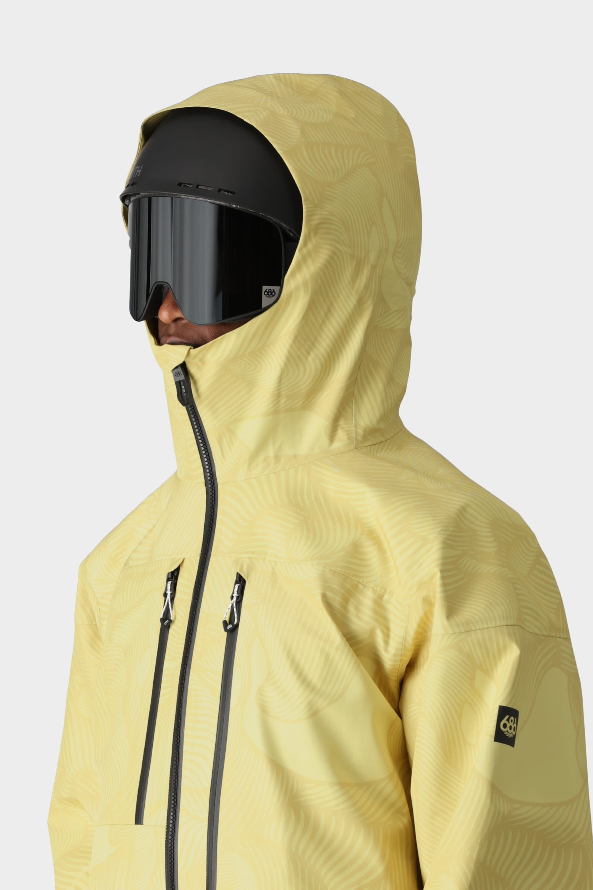 A person wearing the 686 Mens GORE-TEX 3L ATV Jacket in yellow, paired with a black ski helmet and large black goggles from 686, faces left against a plain light background.