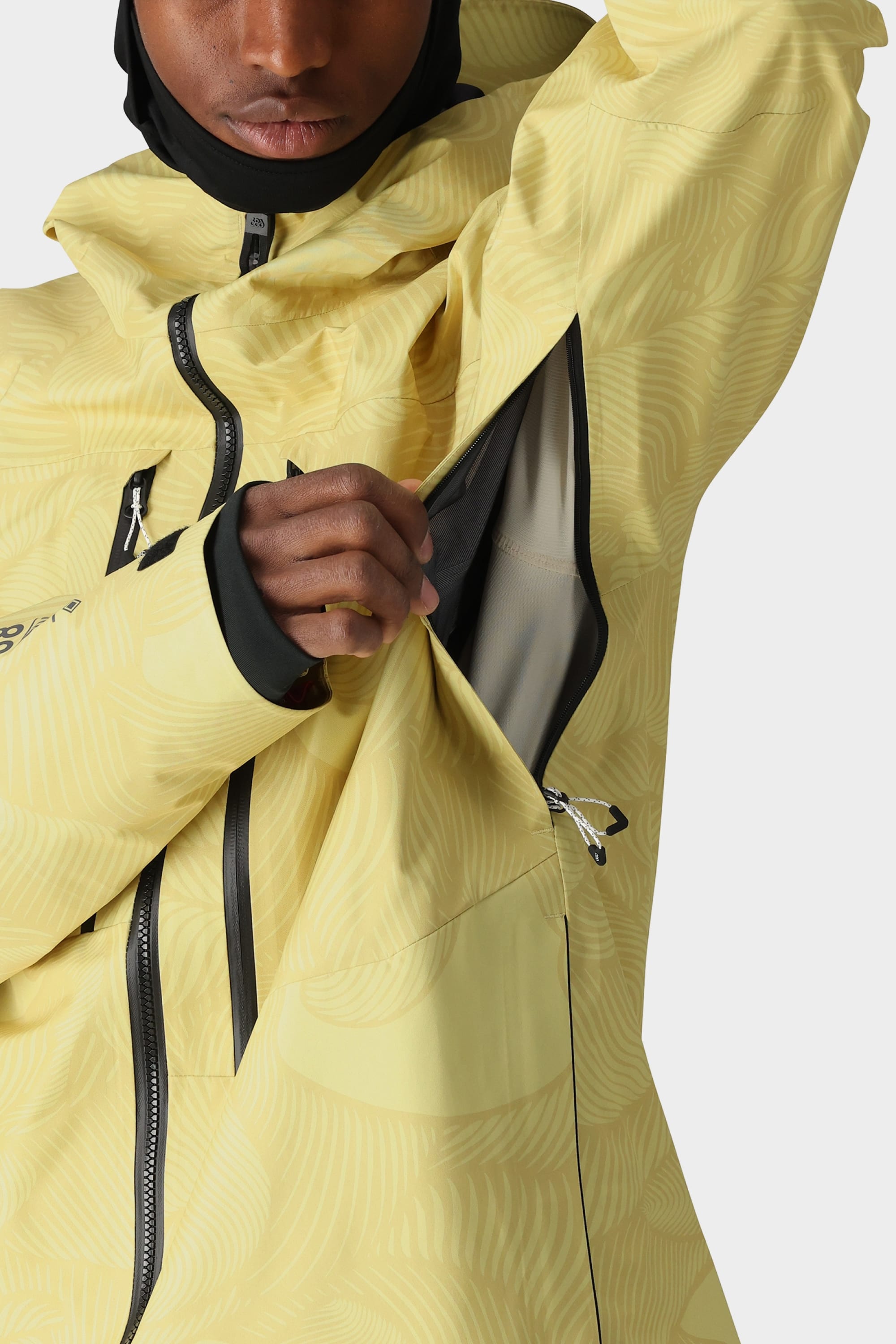 A person wearing the 686 Mens GORE-TEX 3L ATV Jacket made from recycled fabric unzips a vent under their arm, revealing mesh lining. The yellow patterned jacket by 686 features black zippers and is partially shown from the shoulders down.
