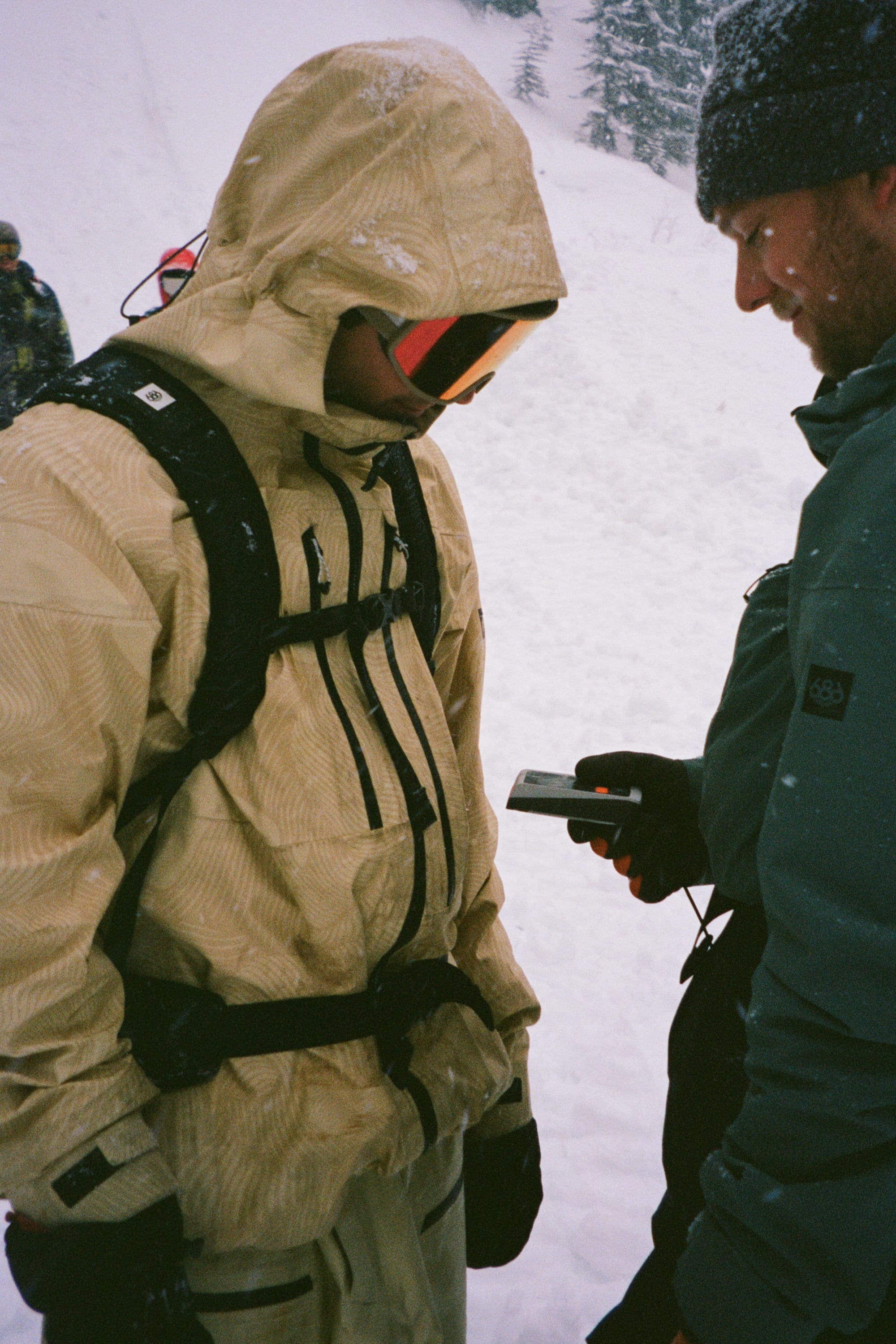 Two people stand in the snow. One, wearing a tan 686 Mens GORE-TEX 3L ATV Jacket and goggles, faces another in a blue jacket and beanie, showing something on a handheld device with snowy trees in the background.