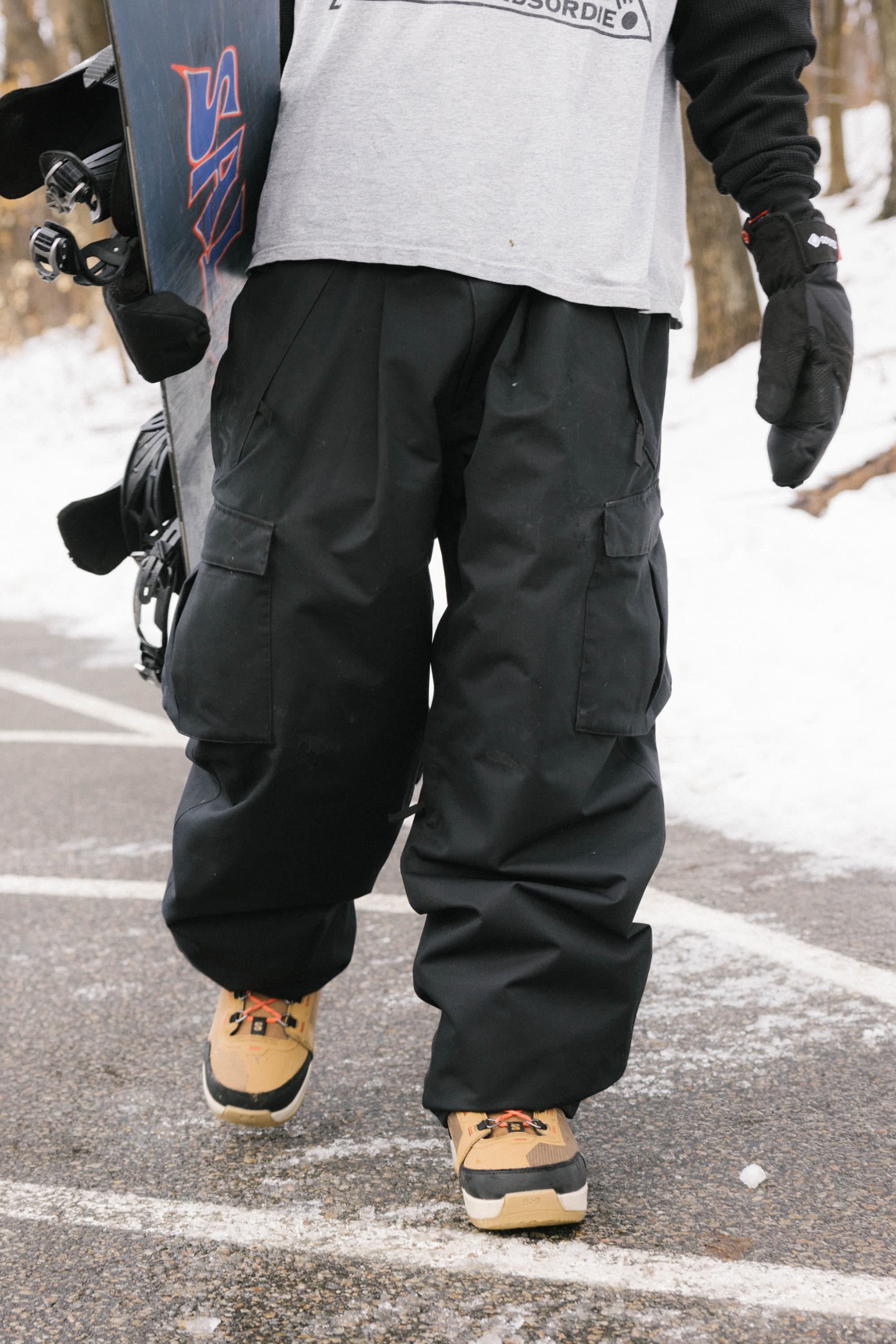 Wearing 686 Men's Essox™ Cargo Pant in black and tan snow boots, a person holds a snowboard while walking on a snowy path in the woods. Their upper body and face are not visible.