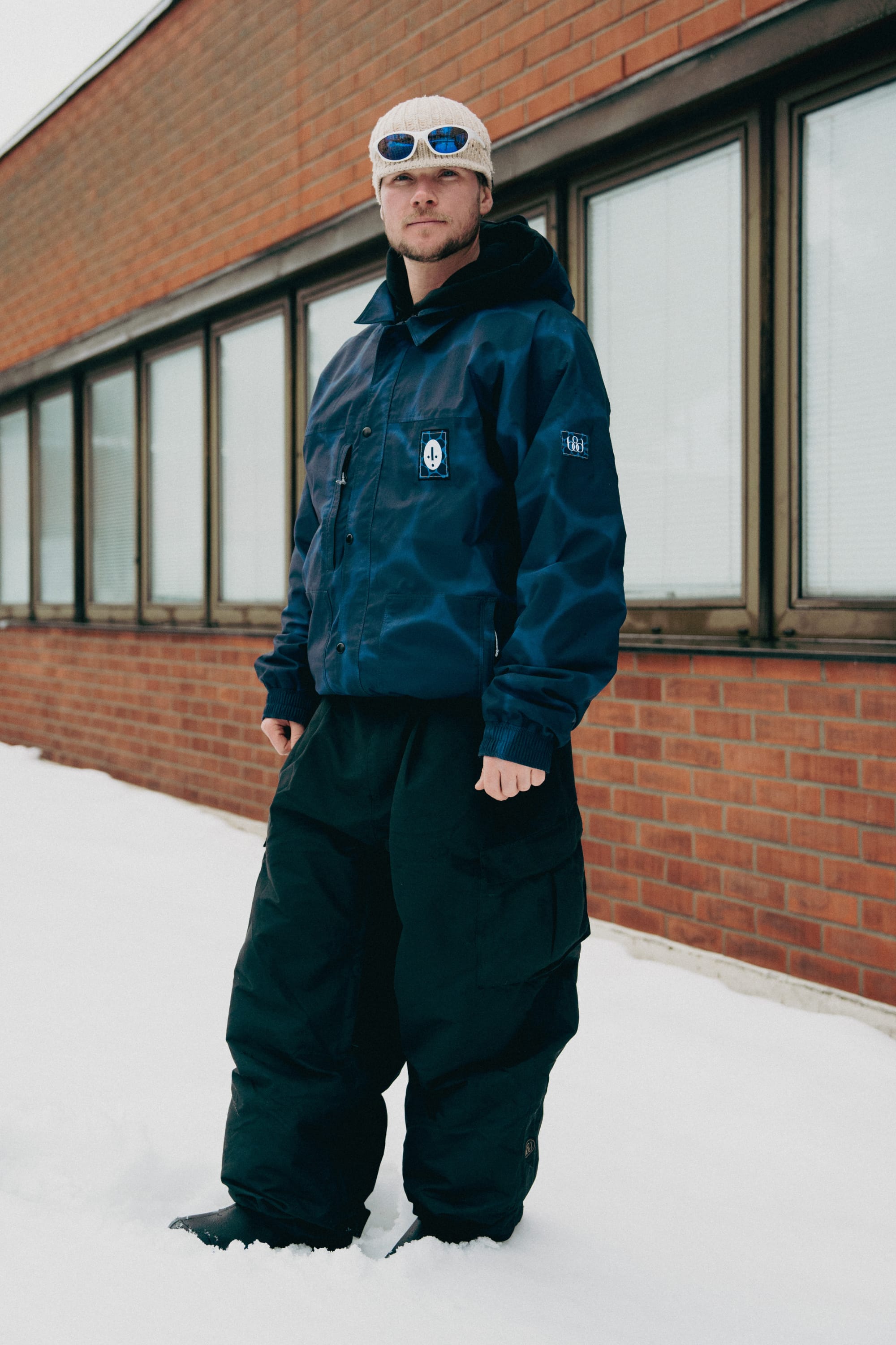 A person stands in snow wearing a blue jacket, 686 Men's Essox™ Cargo Pant by 686, a winter hat, and sunglasses in front of a brick building with large windows.