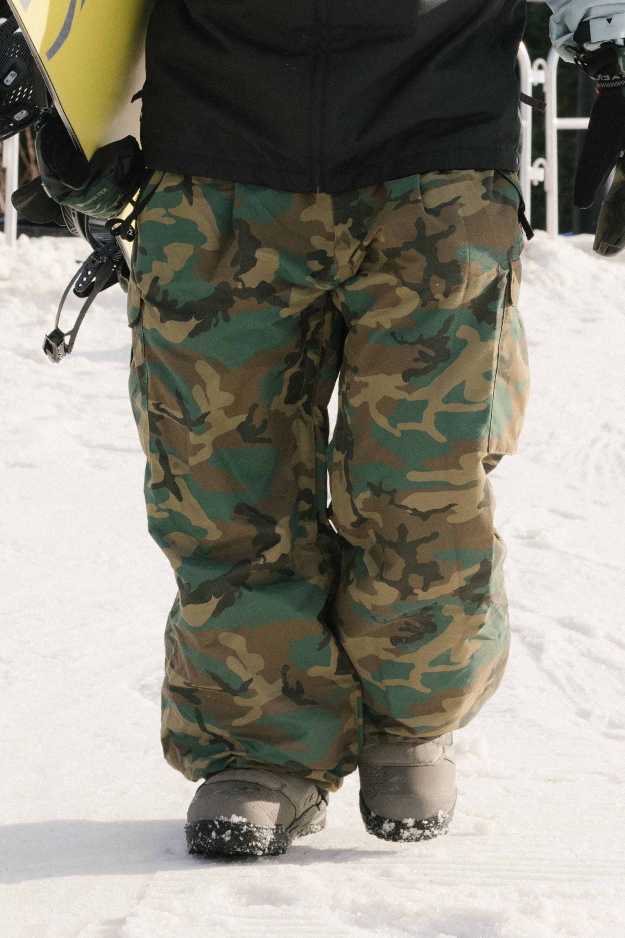 A person in 686 Men's Essox™ Cargo Pant stands on snow, holding a snowboard and helmet, wearing gray boots.