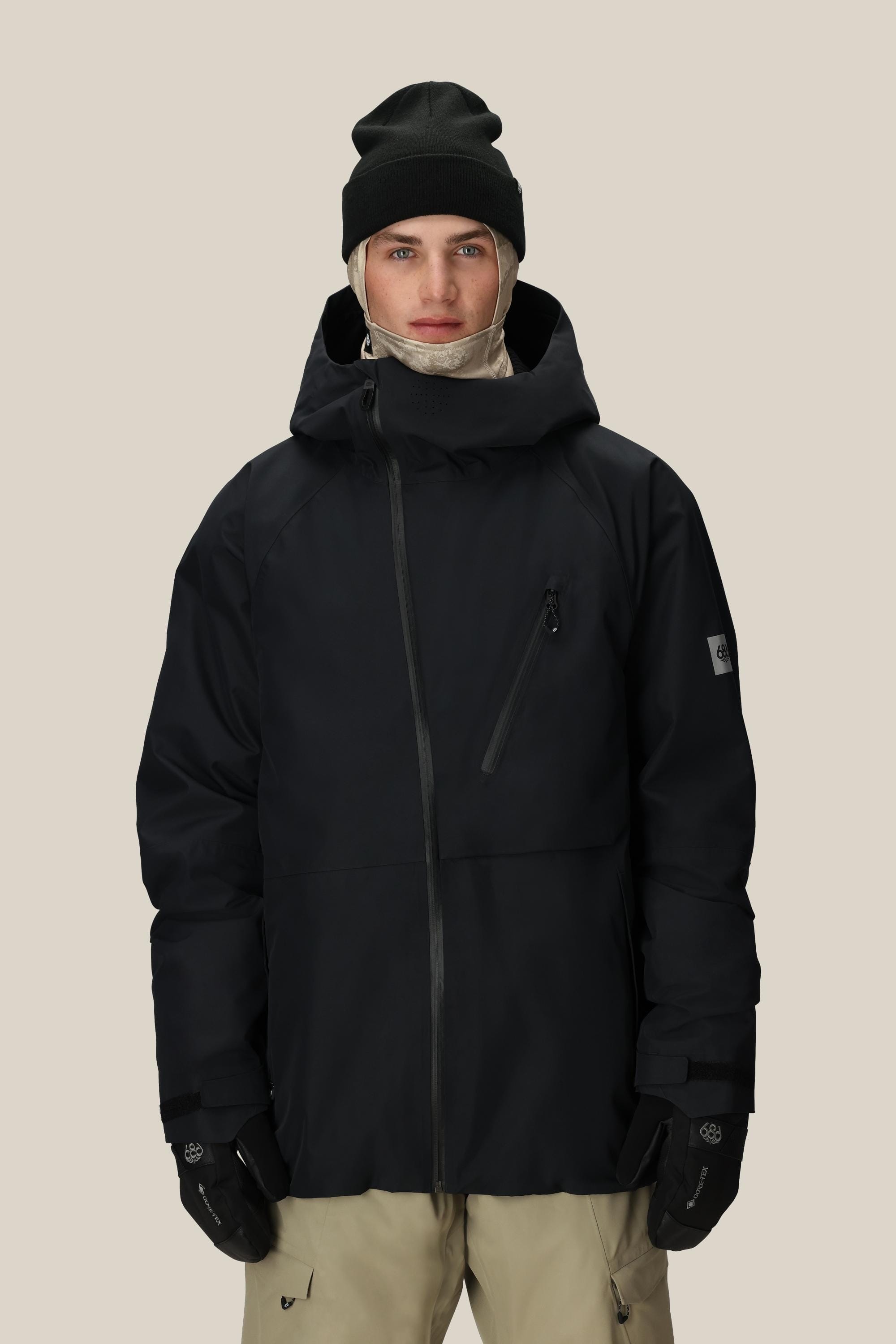 A person wears the 686 Men's Hydra™ Thermagraph® Jacket by 686—a black do-it-all jacket with body-mapped lining and chest zipper pocket—plus a black beanie, beige pants, black gloves, and a beige neck gaiter against a plain beige background.