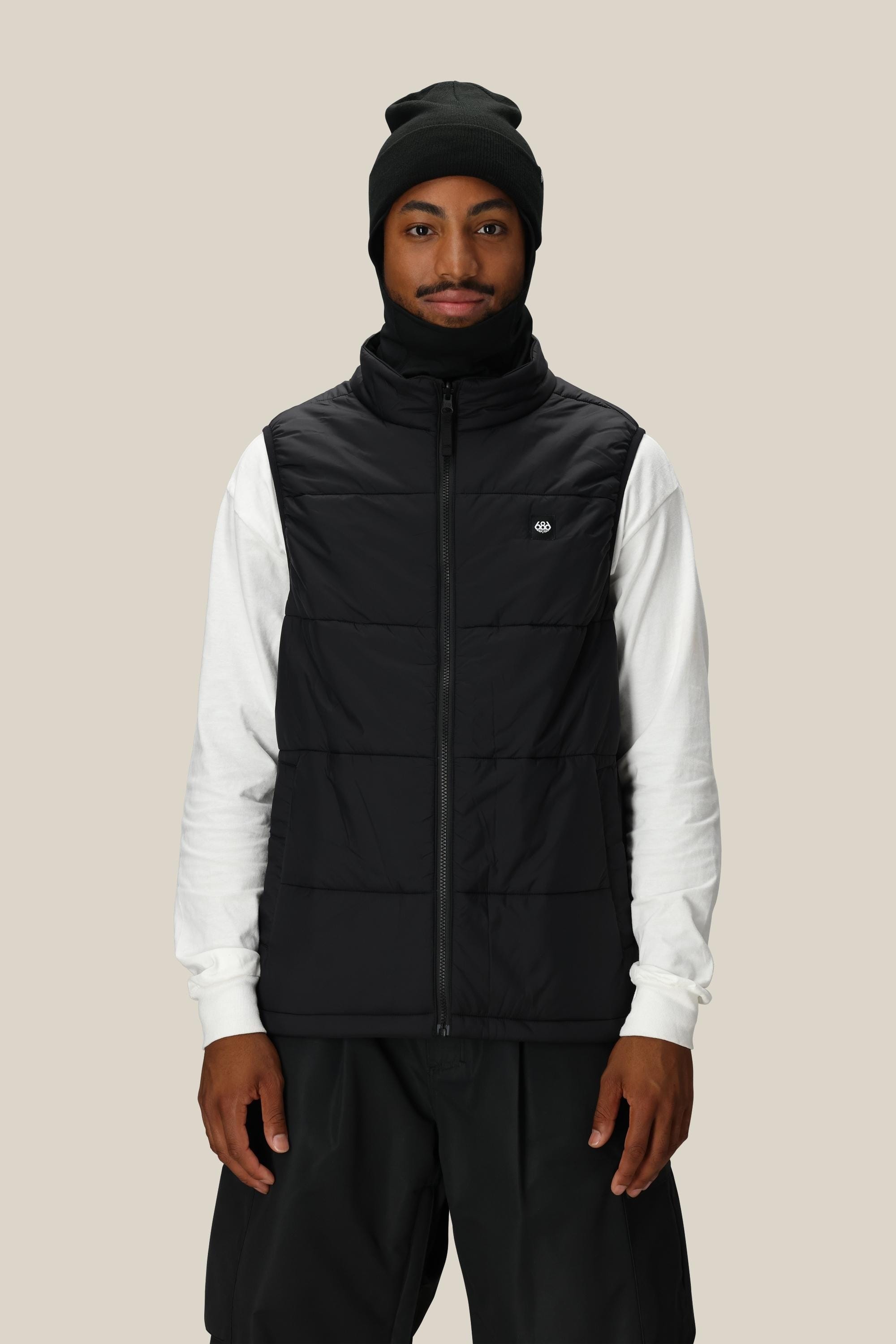 A man wearing the 686 Men's SMARTY® 3-In-1 Rodeo™ 3L Jacket over a white long-sleeve shirt, with a black beanie and black pants, stands facing the camera against a plain beige background.