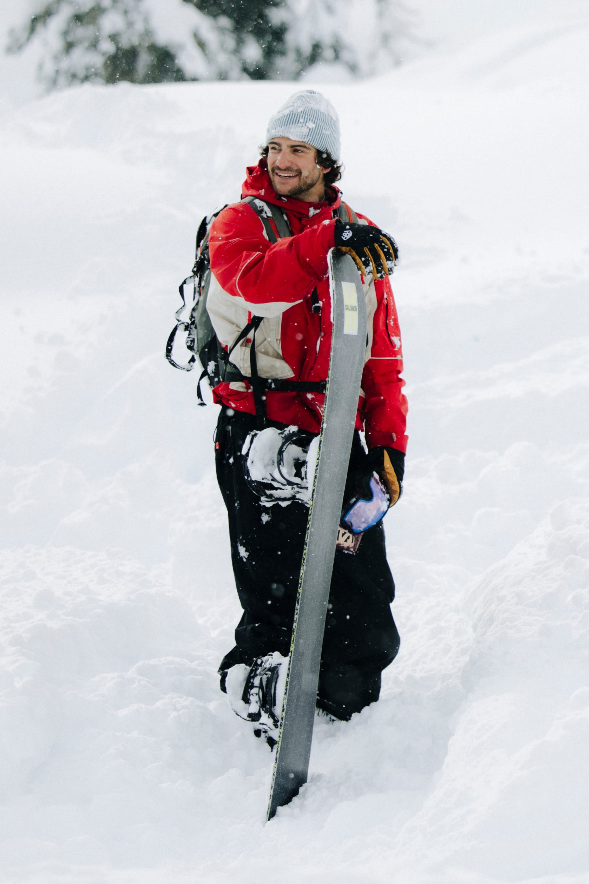 A person in a 686 Men's 2002 X3 3L Shell Jacket and gray beanie stands smiling in deep snow, holding a snowboard upright with snowy trees in the background.