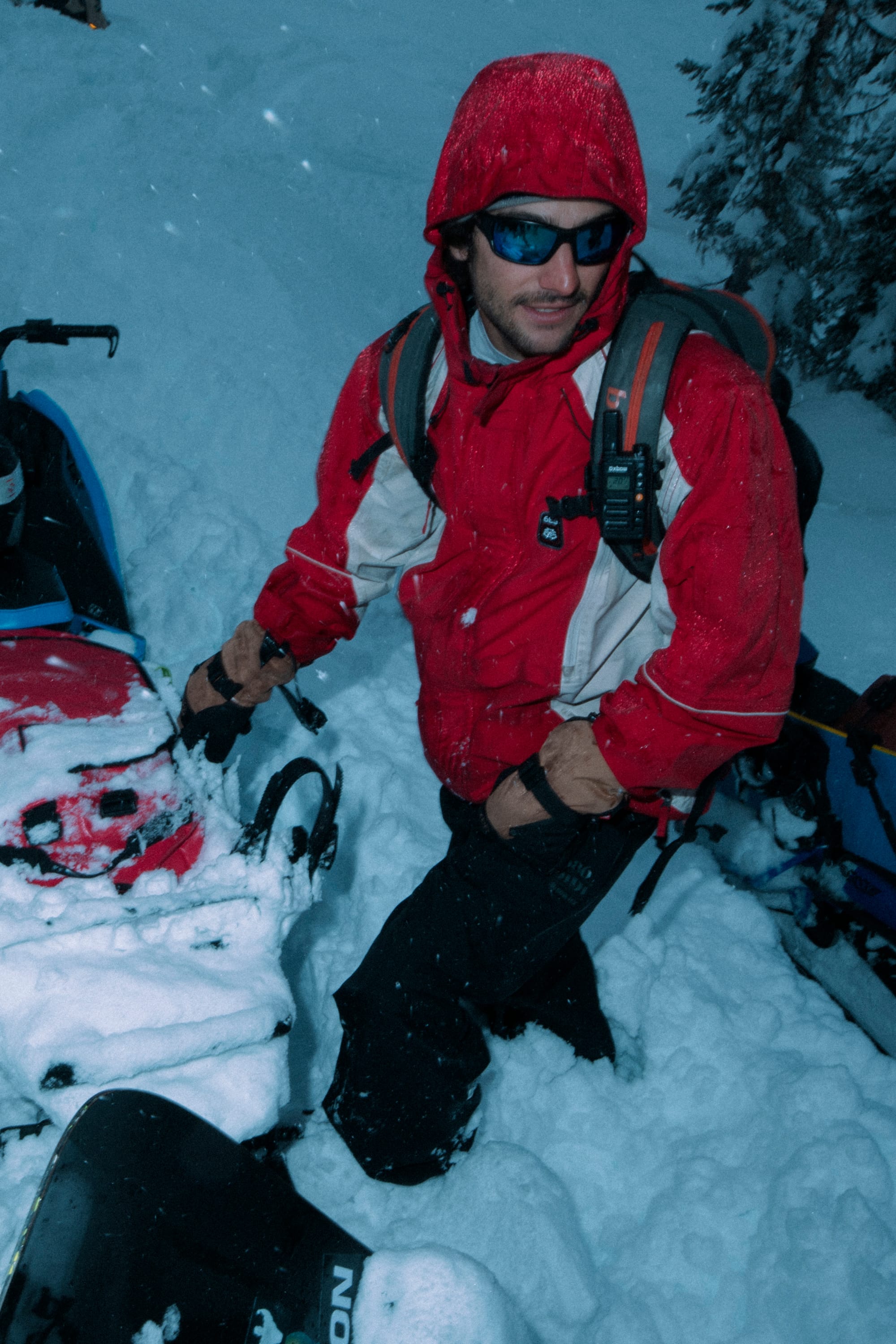 A person wearing the 686 Men's 2002 X3 3L Shell Jacket in red, plus sunglasses and gloves, stands in deep snow next to a snowmobile, with trees and snowy terrain behind.