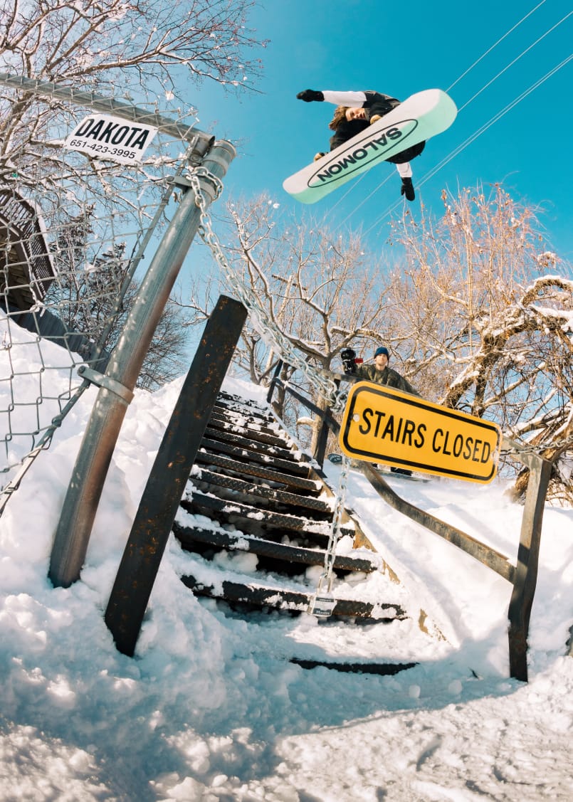 A snowboarder jumps over a snow-covered staircase with a “STAIRS CLOSED” sign, while another person watches below. Bare trees and a bright blue sky form the background. A chain blocks the stairs.