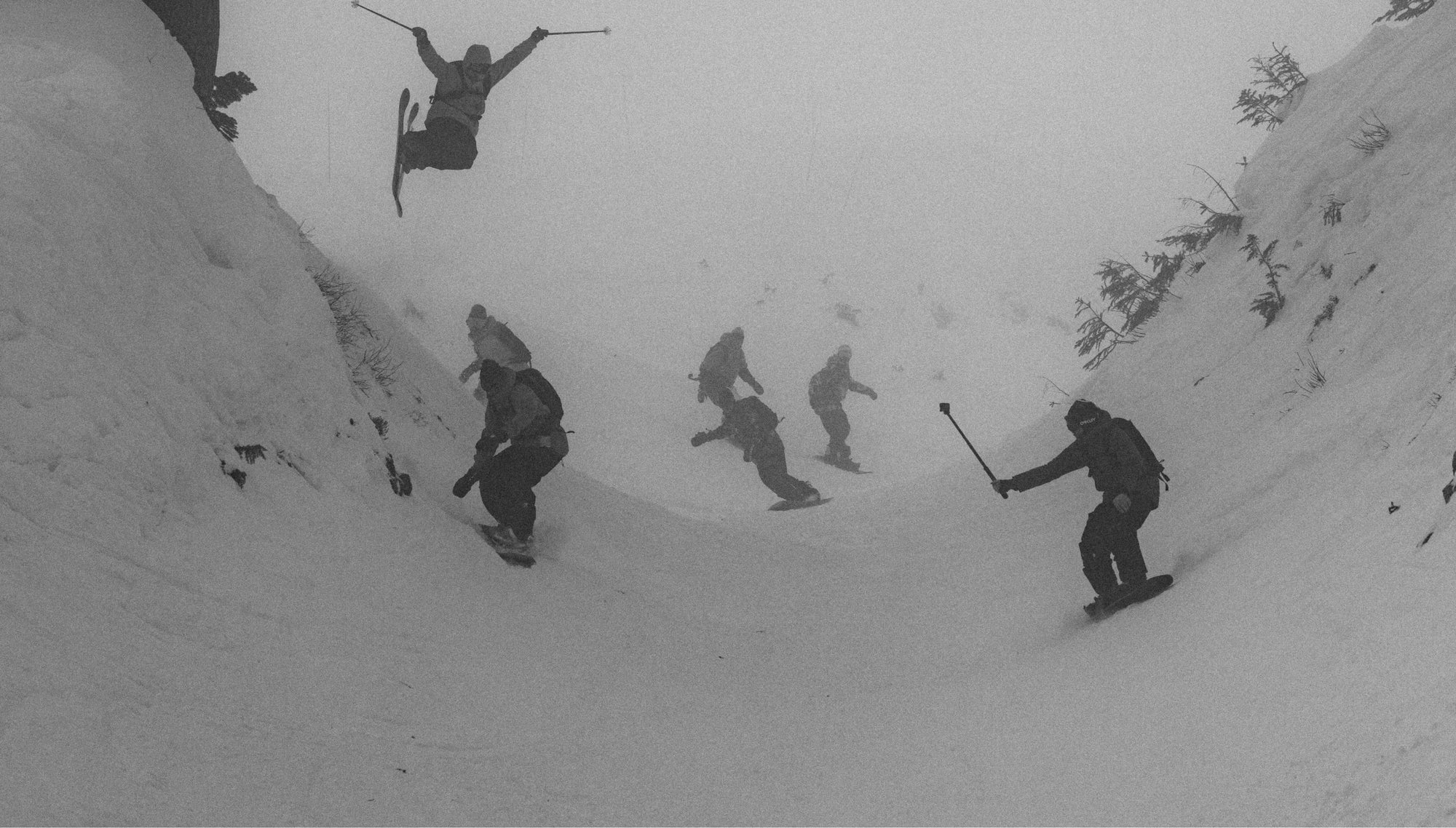 A group of skiers and snowboarders ride down a snowy slope in foggy conditions; one skier is captured mid-air with arms and legs spread. The scene is in black and white.
