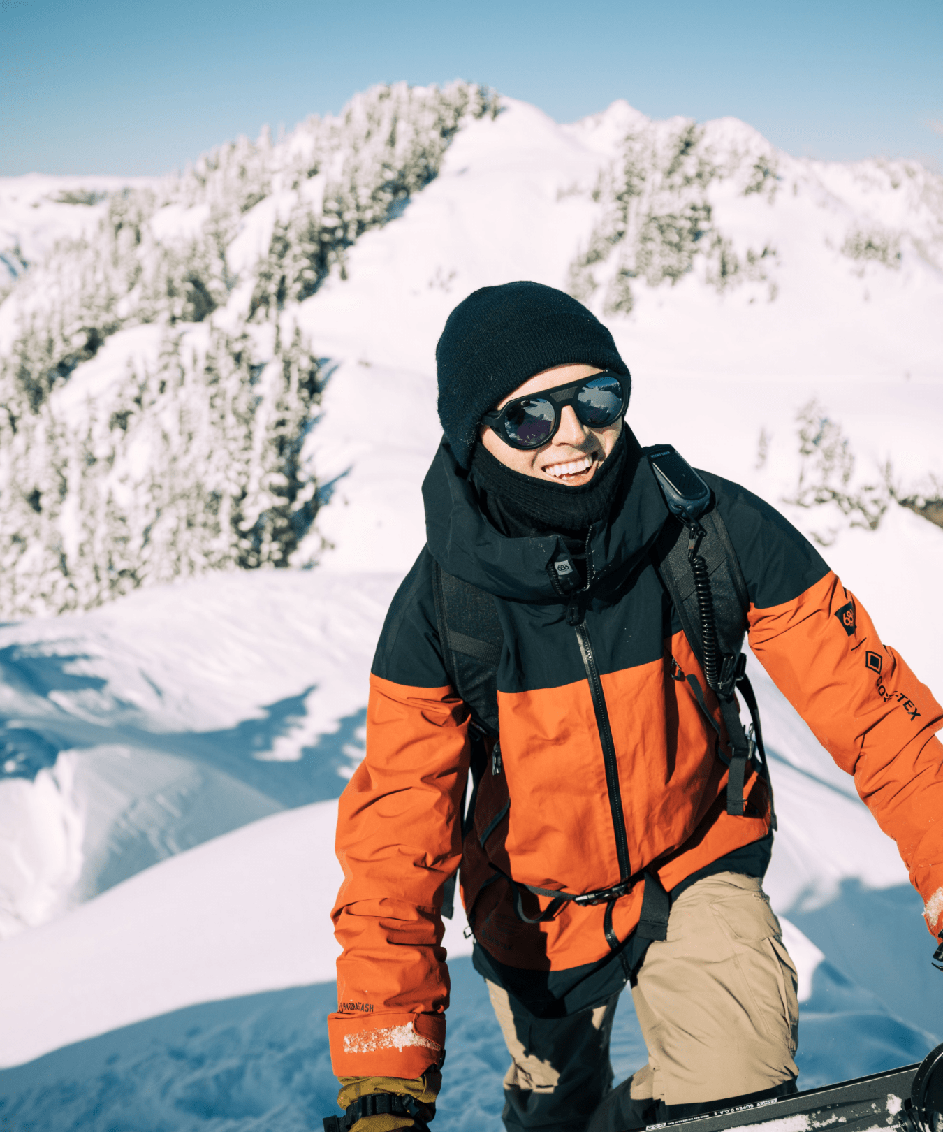 A person in a red and black jacket, tan pants, gloves, hat, and sunglasses smiles while standing in a snowy mountain landscape under a clear blue sky. Snow-covered trees and peaks are in the background.