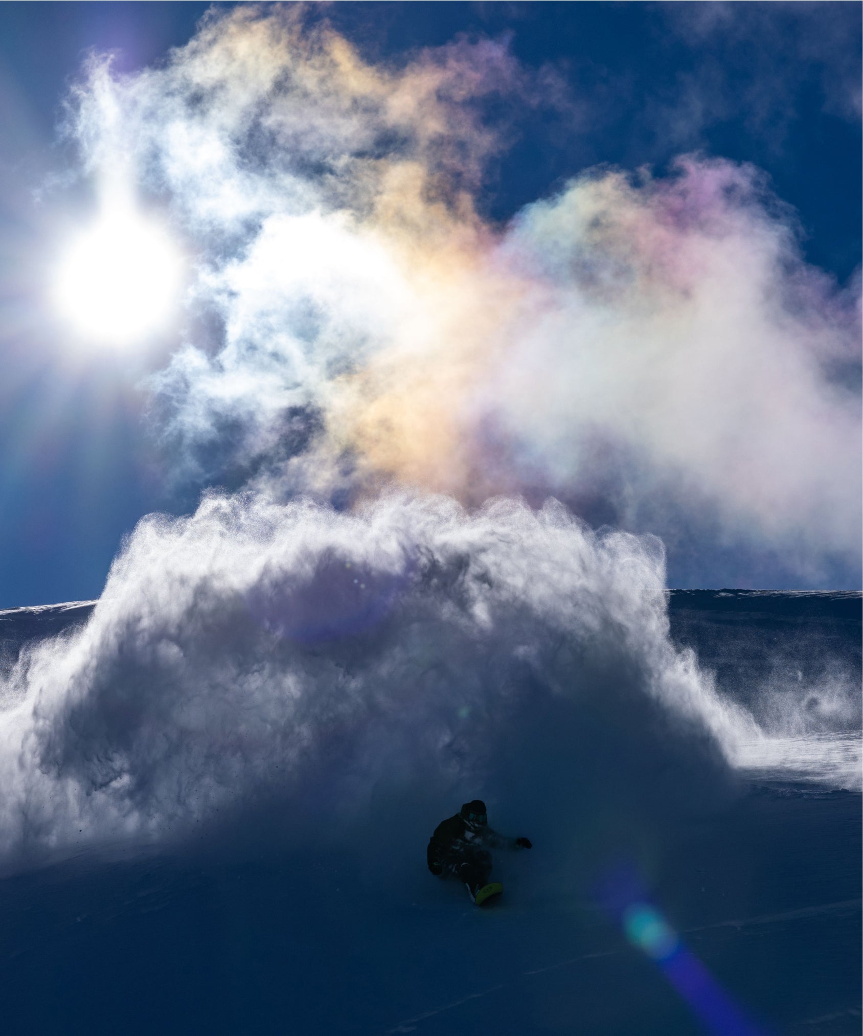 A snowboarder pictured as he rides through clouds of snow produced by the powder on the mountain, backlit by the sun.