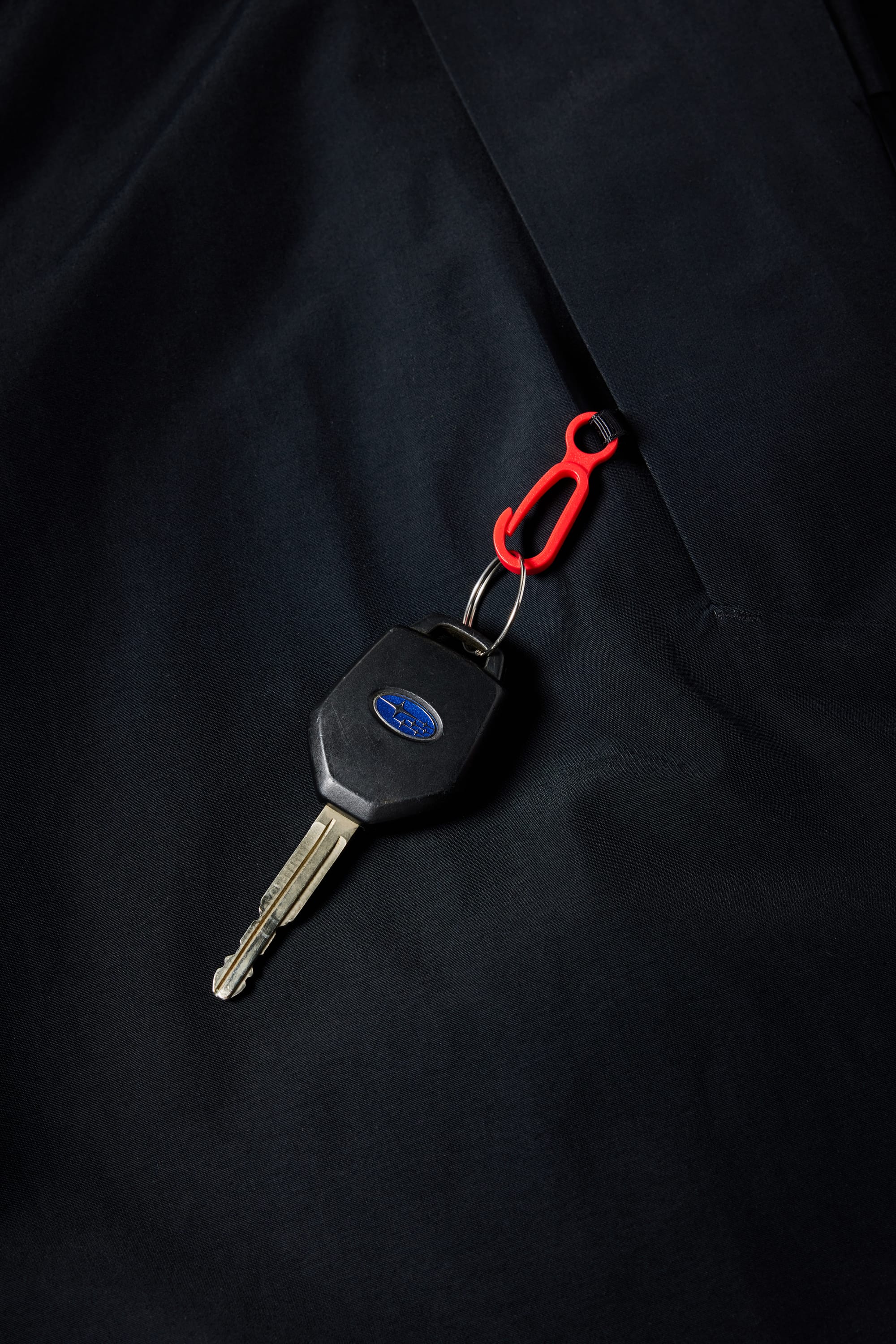 A car key with a black plastic head and blue oval logo is attached to a small red carabiner, resting on the pocket of the 686 Girls Athena Insulated Jacket by 686, set against a dark fabric surface.