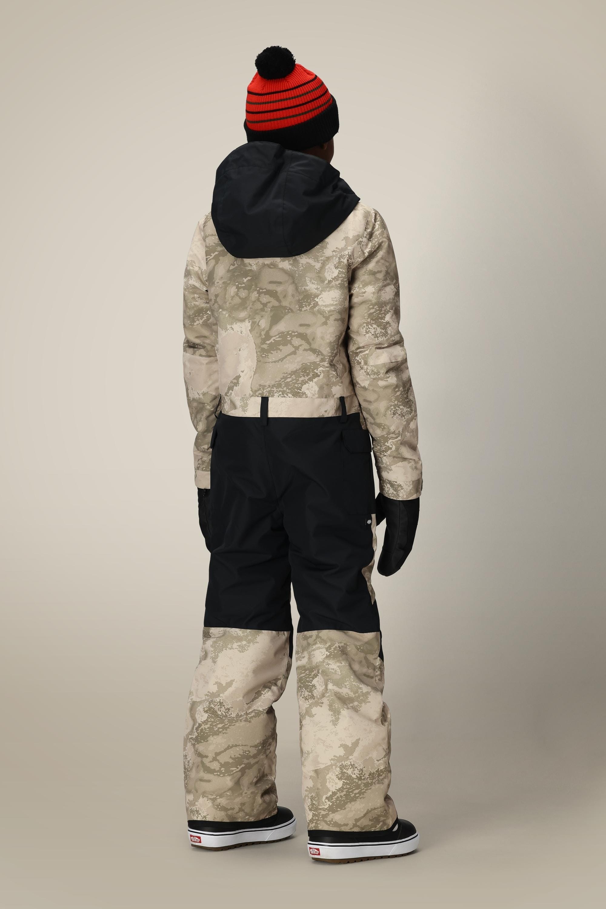 A person faces away, wearing the 686 Boys Shazam Insulated One-Piece in tan and black camo by 686, paired with black mittens, boots, and a red-and-black striped pom-pom beanie, set against a plain background.