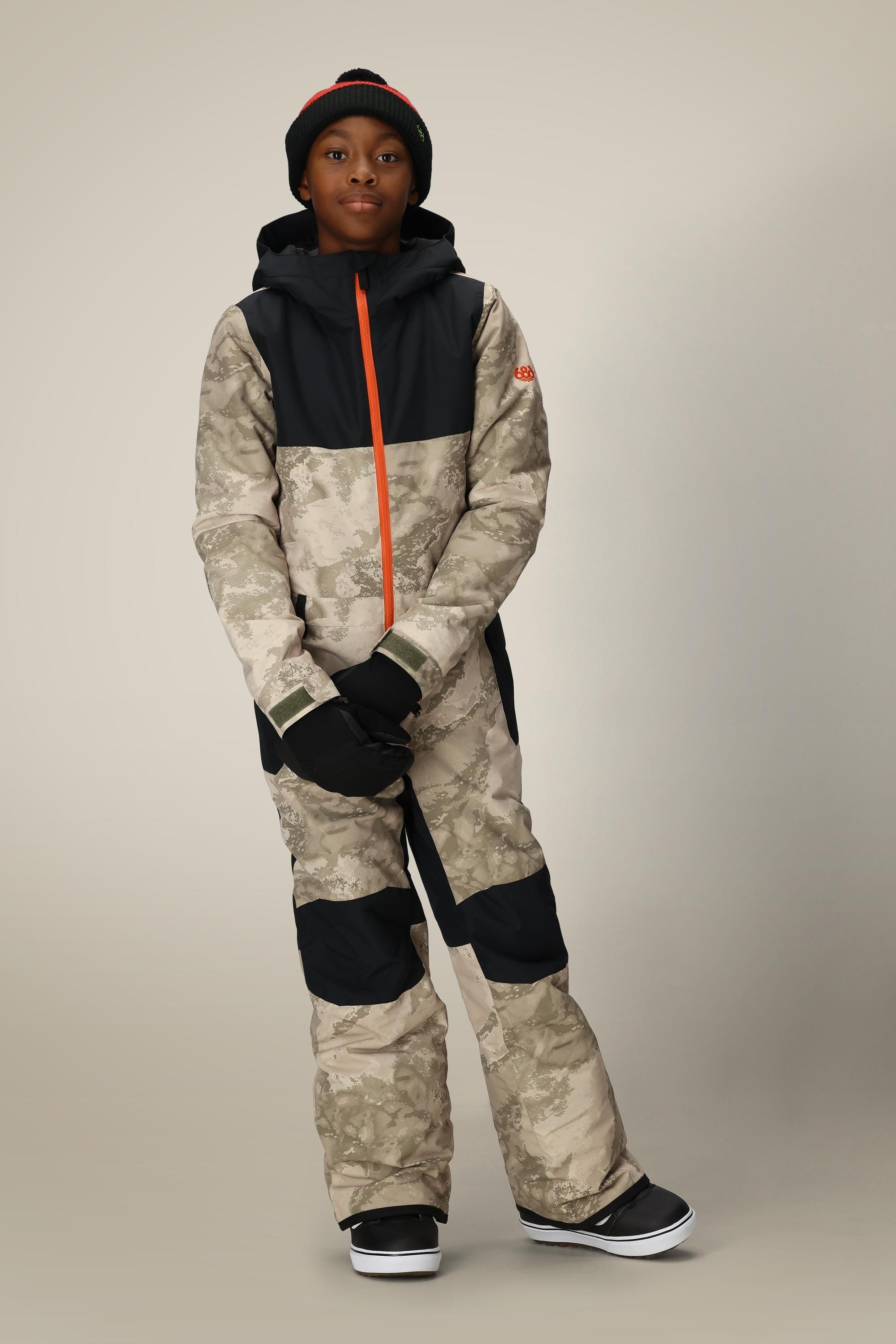 A child wears the 686 Boys Shazam Insulated One-Piece by 686—a camo-patterned snowsuit with black panels and orange zipper—styled with black gloves, beanie, and snow boots against a plain beige background.