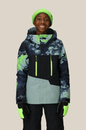 A smiling child wears the 686 Boys Geo Insulated Jacket, featuring green and black geometric accents, paired with a neon green beanie, black snow pants, and neon gloves, standing against a plain beige background. Brand: 686.