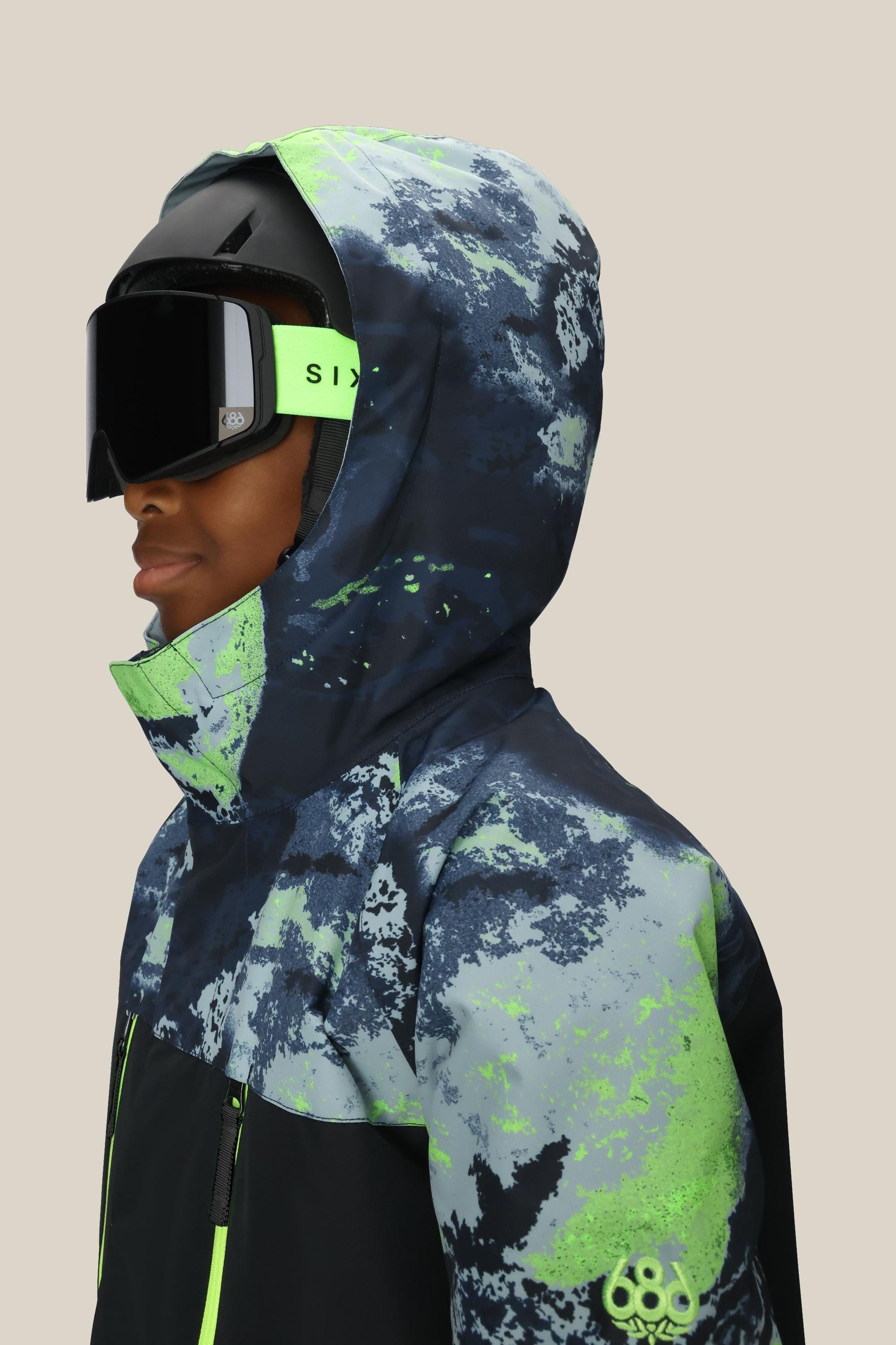 A person is shown from the side against a plain beige background wearing a black helmet, large black ski goggles, and the 686 Boys Geo Insulated Jacket by 686, which has a blue, green, and white abstract geometric pattern.