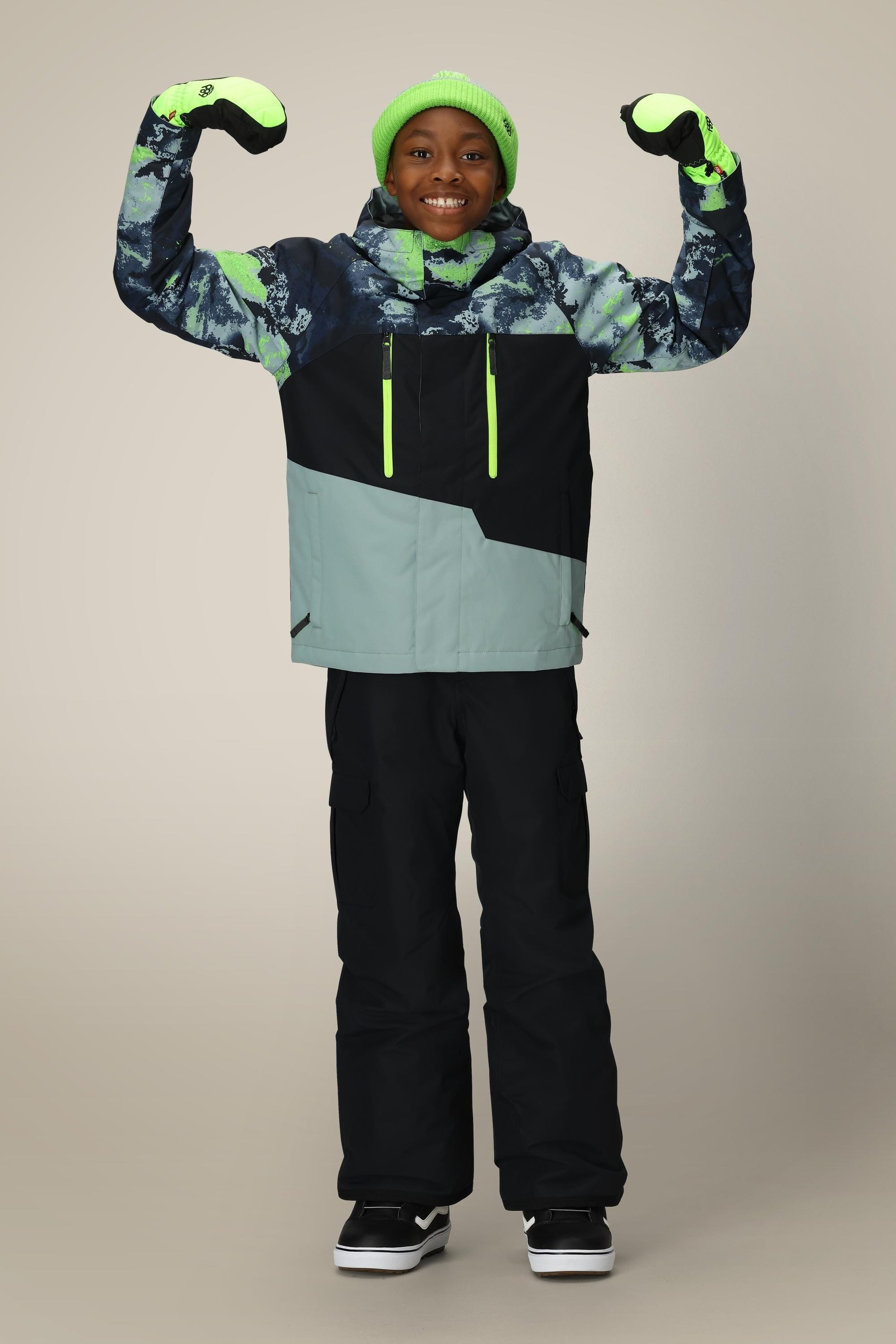 A smiling child wears the 686 Boys Geo Insulated Jacket by 686 with neon green accents, a green beanie, black snow pants, and gloves, posing with arms flexed against a neutral background.