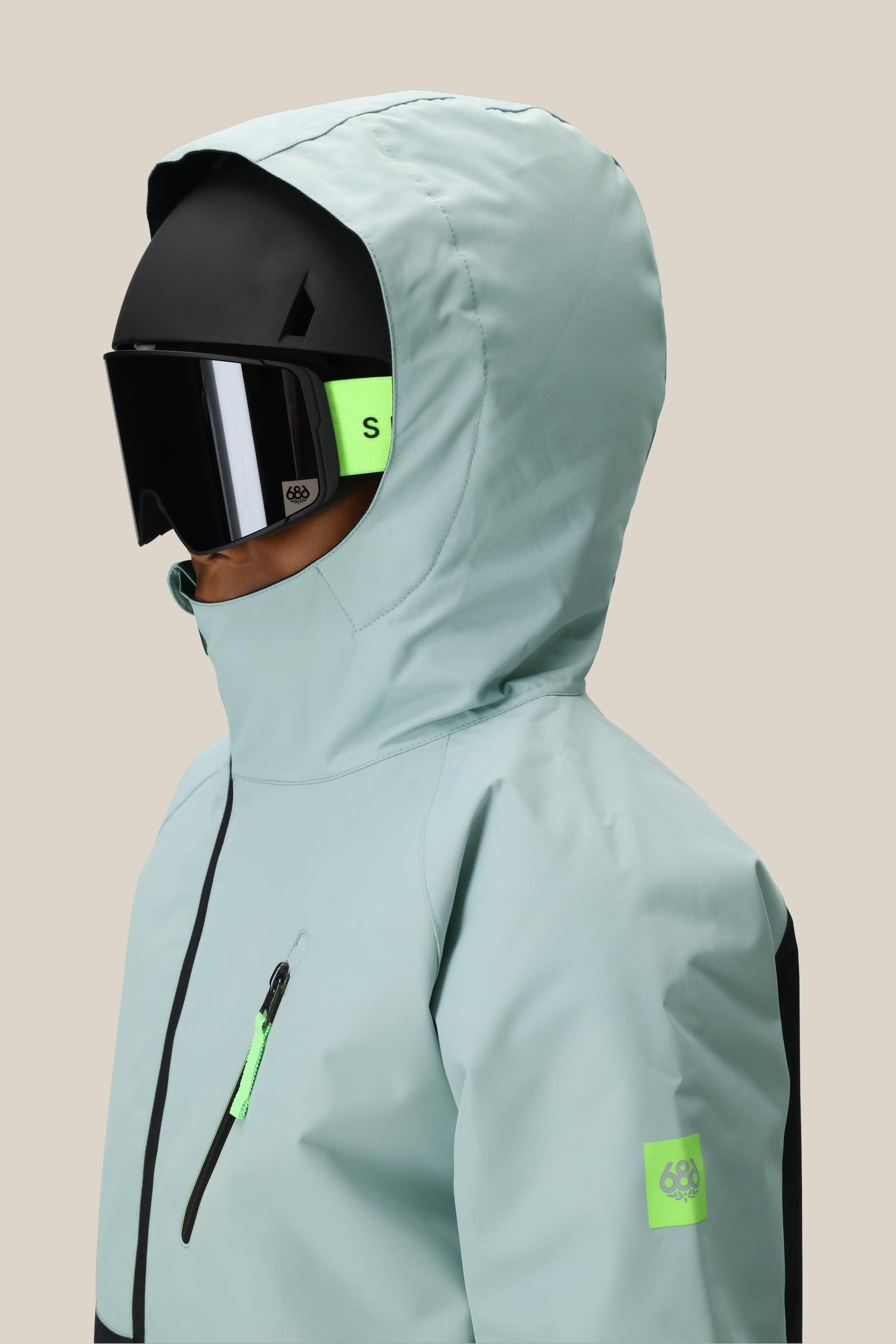 A boy wears the 686 Boys Hydra Insulated Jacket in light blue, paired with a black ski helmet and large black ski goggles with a neon green strap, shown from the side against a plain beige background.