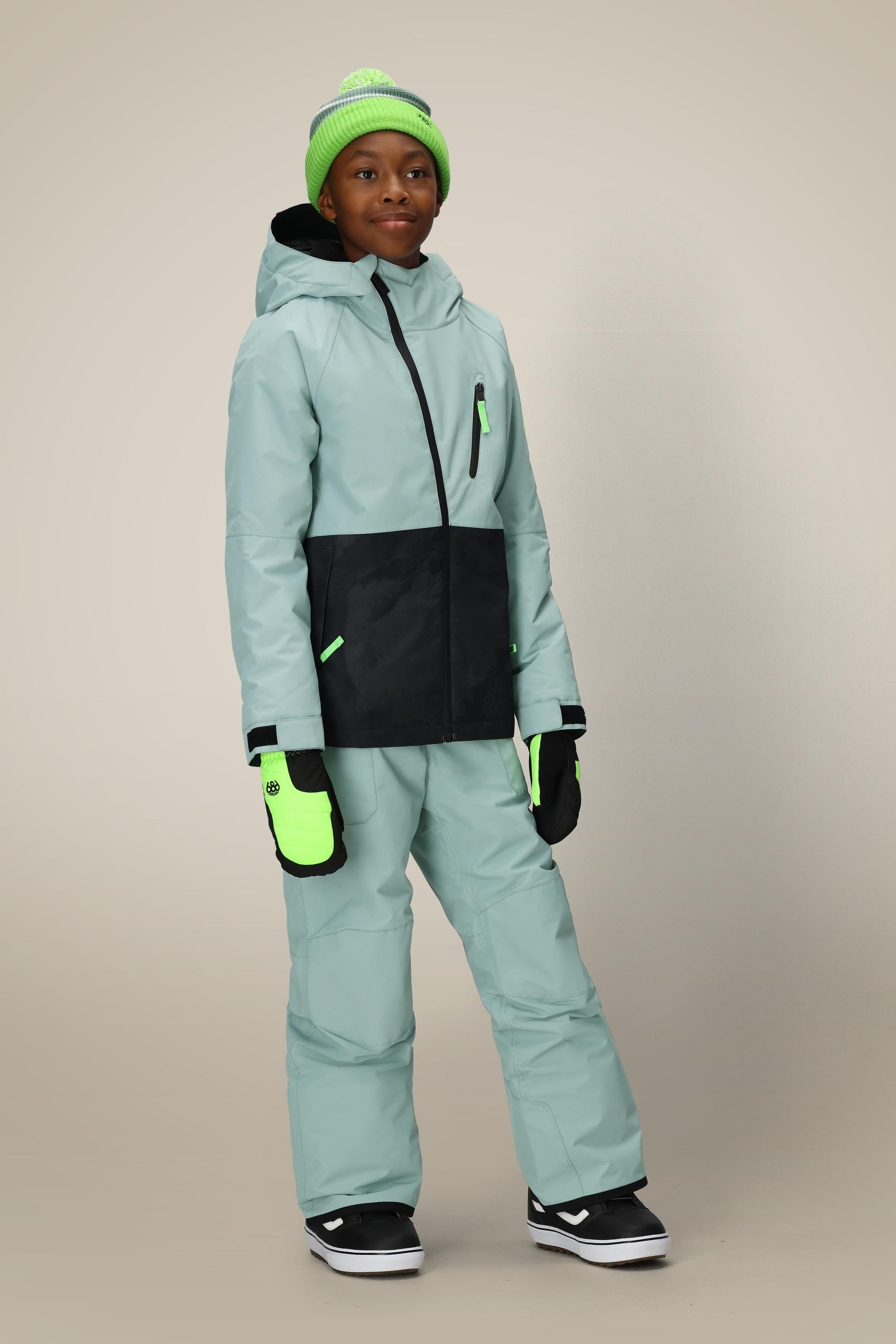 A young person stands facing forward wearing the 686 Boys Hydra Insulated Jacket in light green and black, matching snow pants, neon green mittens, a green pom-pom beanie, and black-and-white snow boots.