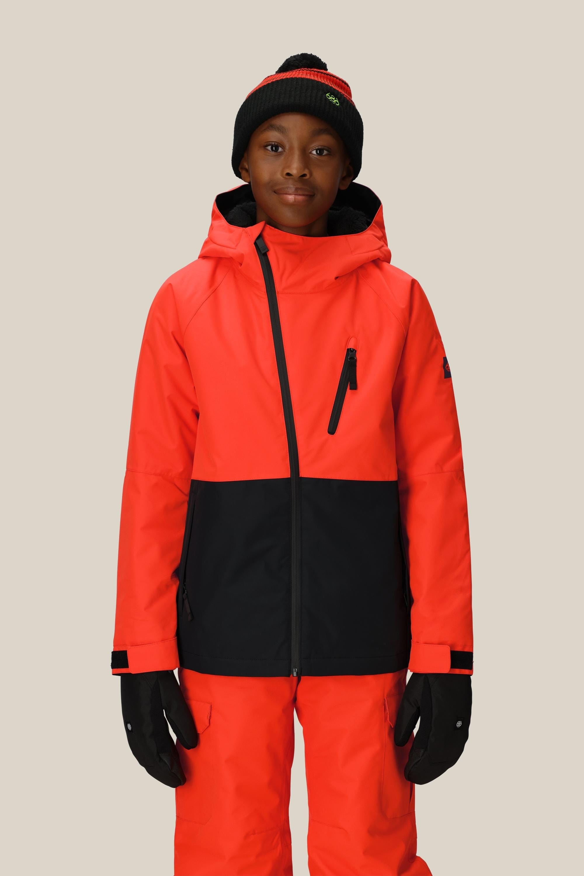 A young person faces forward in a 686 Boys Hydra Insulated Jacket, bright orange and black with body-mapped lining, matching pants, black gloves, and a black beanie with a red stripe, set against a plain beige background.