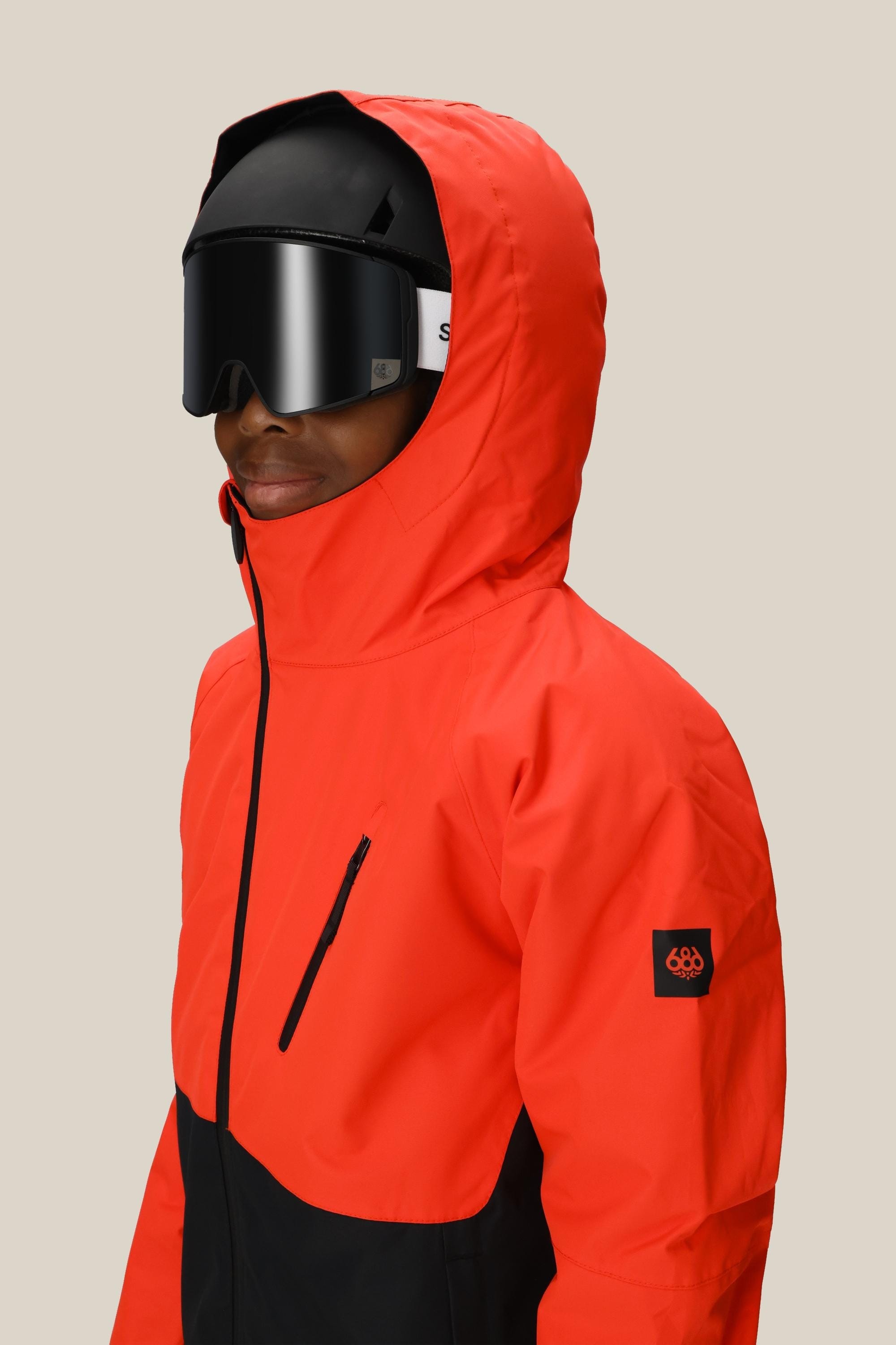 A boy wearing the 686 Boys Hydra Insulated Jacket in bright red and black, paired with black ski goggles and a helmet, stands against a plain beige background.