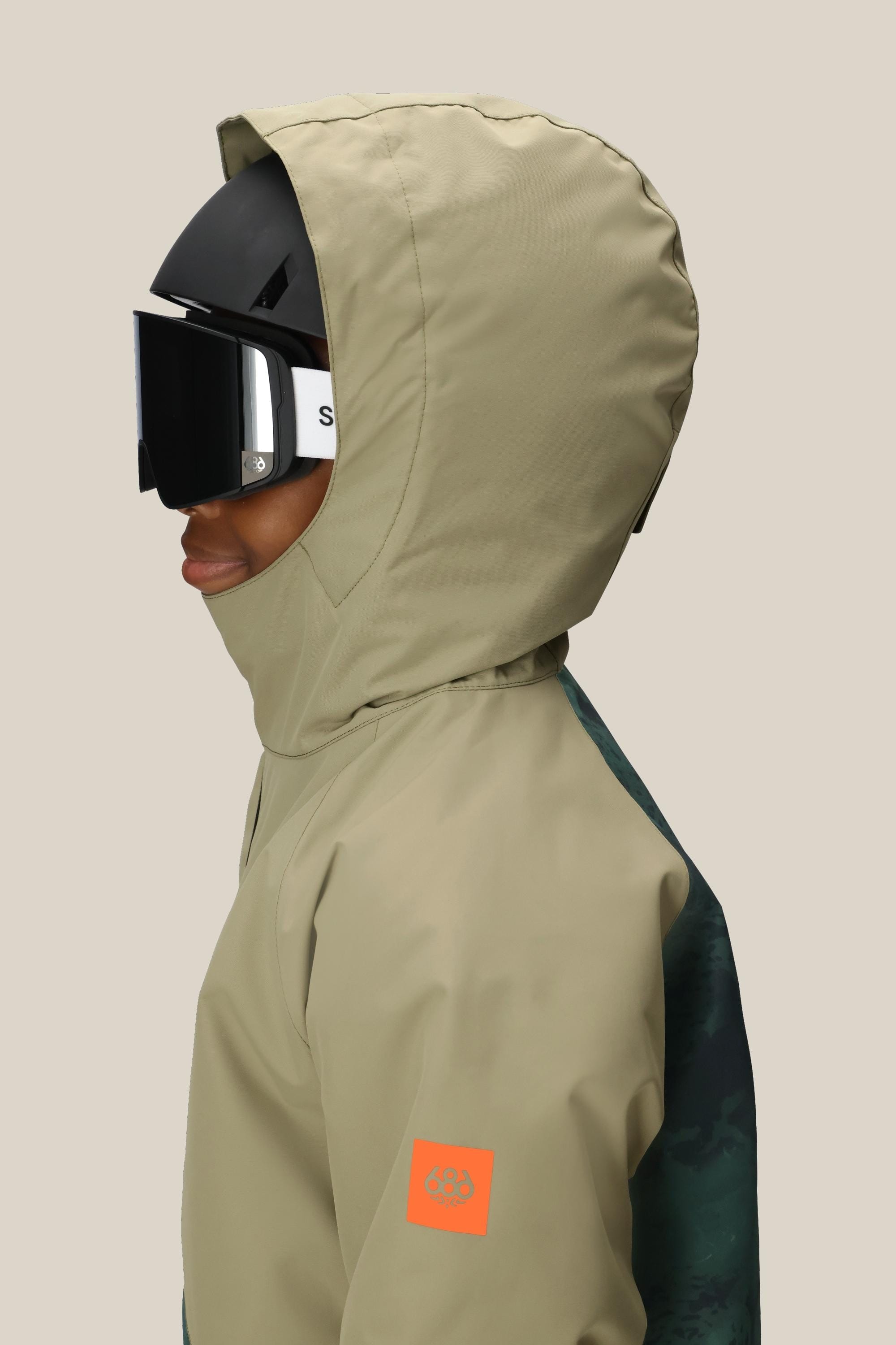 A boy wears the 686 Boys Hydra Insulated Jacket in beige with an orange sleeve logo patch, paired with a black helmet and ski goggles, shown in profile against a plain light background. Brand: 686.