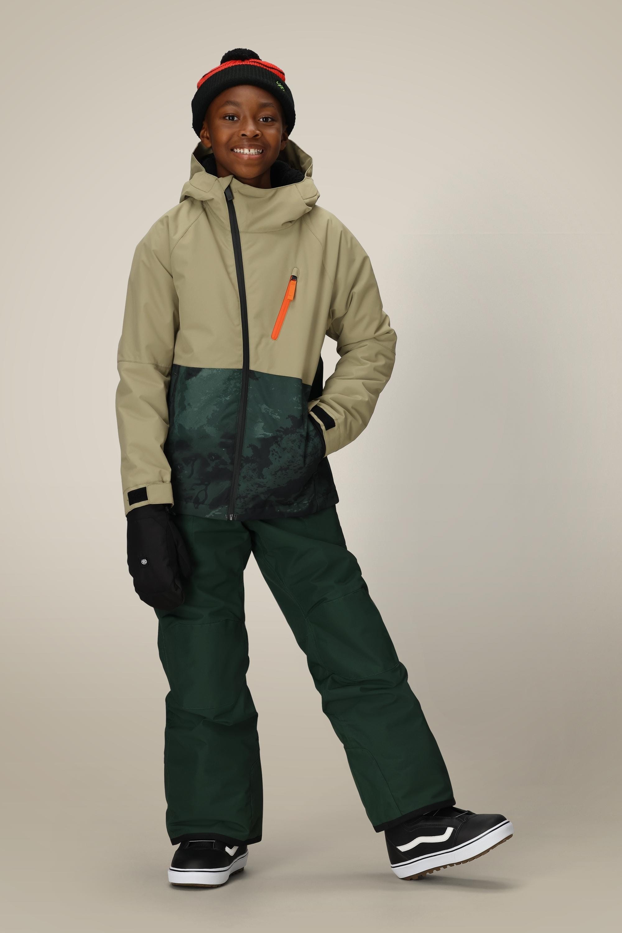 A smiling child wears the 686 Boys Hydra Insulated Jacket, dark green snow pants, black gloves, a black beanie with a red stripe, and black sneakers with white soles against a plain light background.