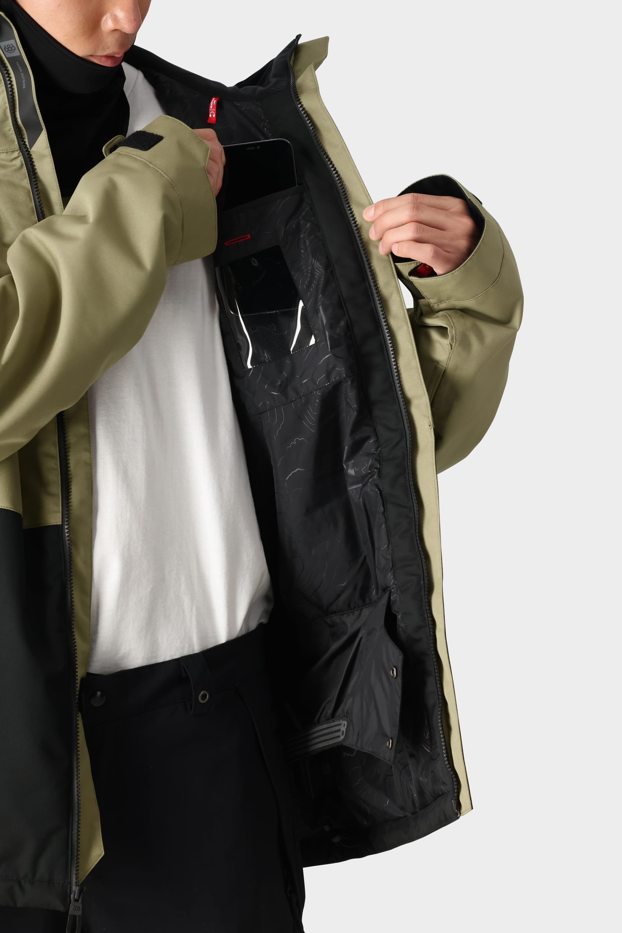 A person wearing the 686 Mens Foundation Insulated Jacket in two-tone green and black opens it over a white shirt to place a smartphone in the inner pocket, ready for their next mountain adventure.