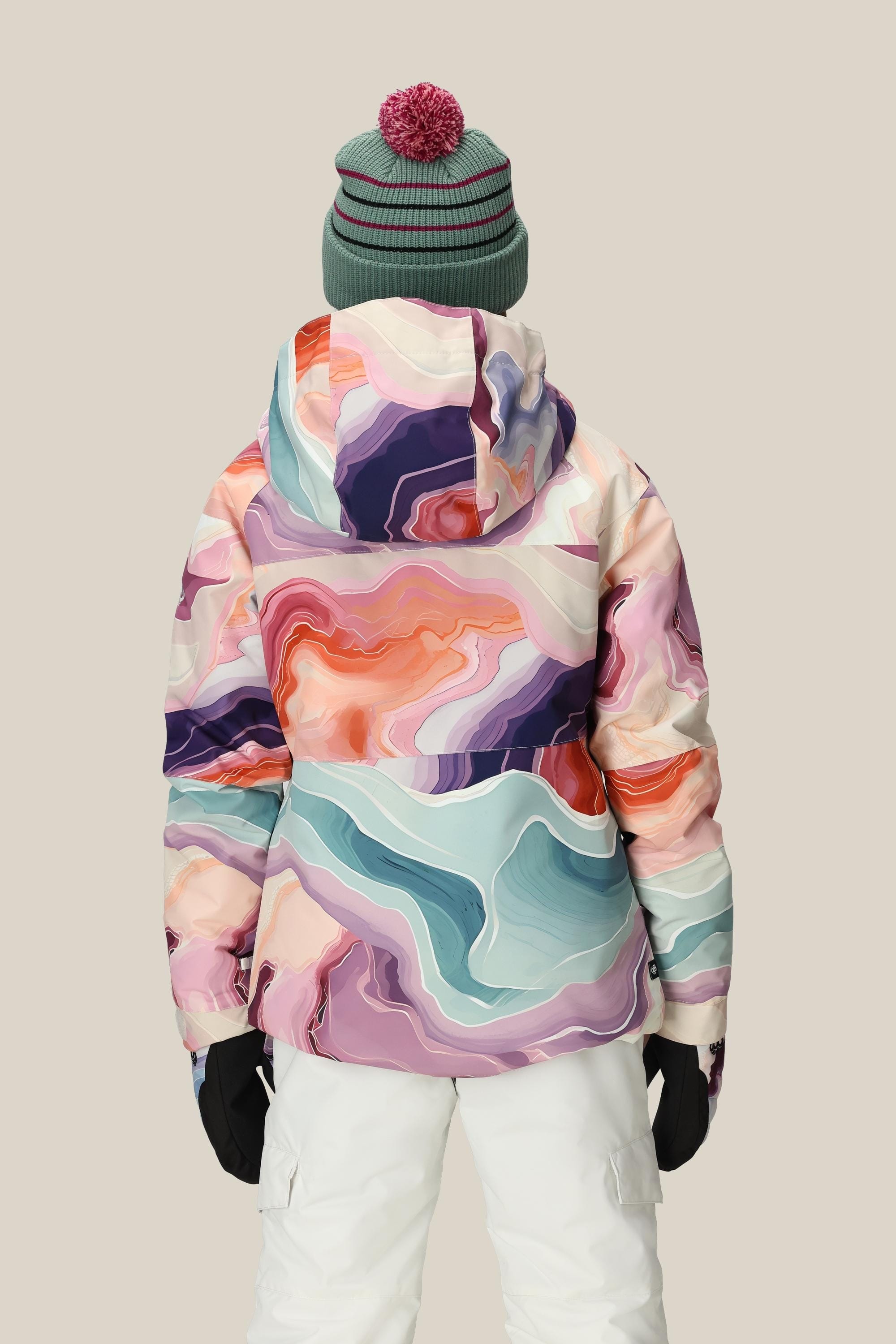 Someone is seen from behind wearing the 686 Girls Athena Insulated Jacket by 686, featuring a colorful abstract pattern. They also wear a green pom-pom beanie, white pants, and black gloves, standing before a plain beige background.