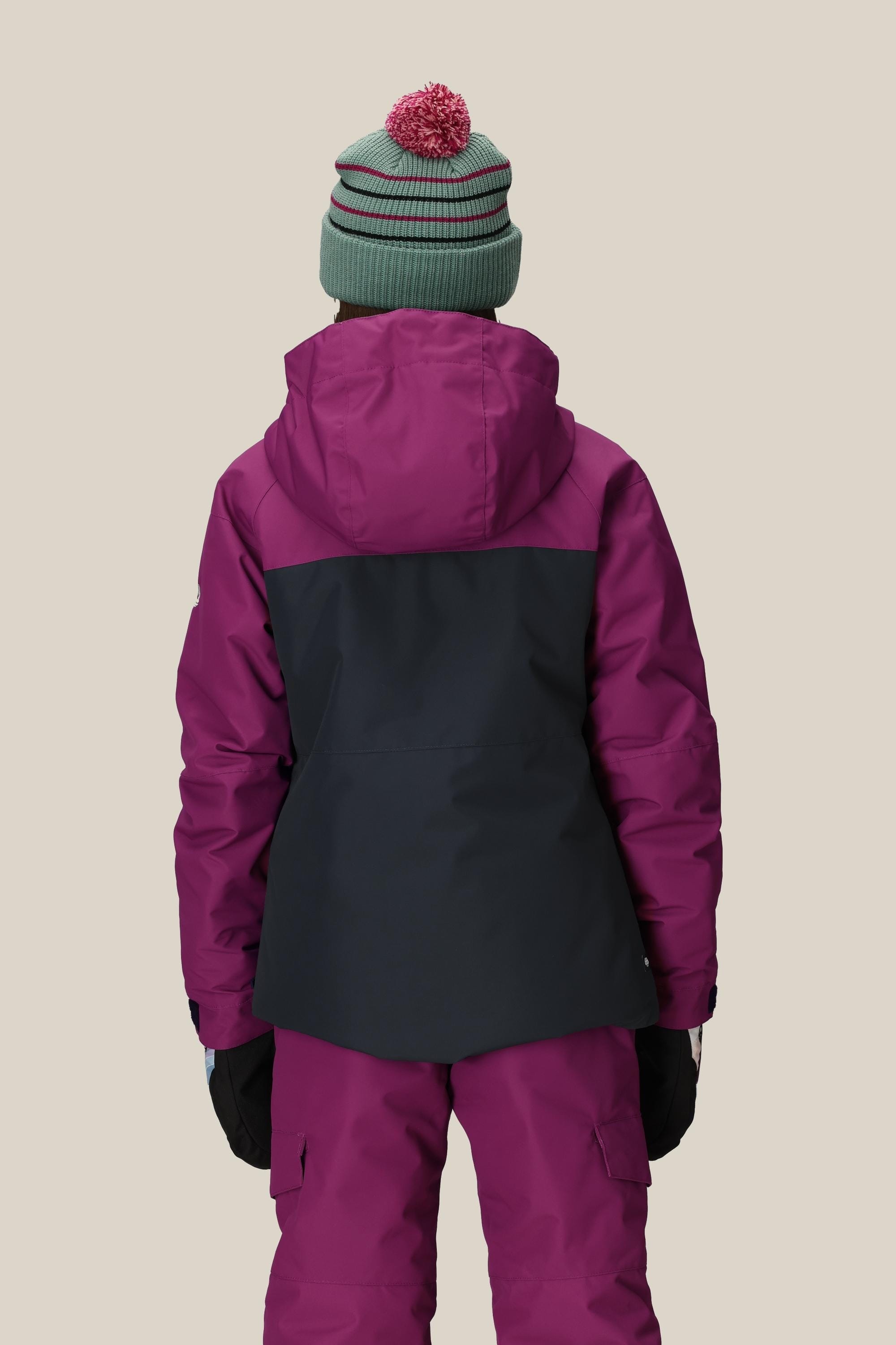 A person is shown from behind against a plain background wearing the 686 Girls Athena Insulated Jacket by 686 in magenta and dark gray, with matching magenta snow pants, black gloves, and a green beanie with red stripes and pom-pom.