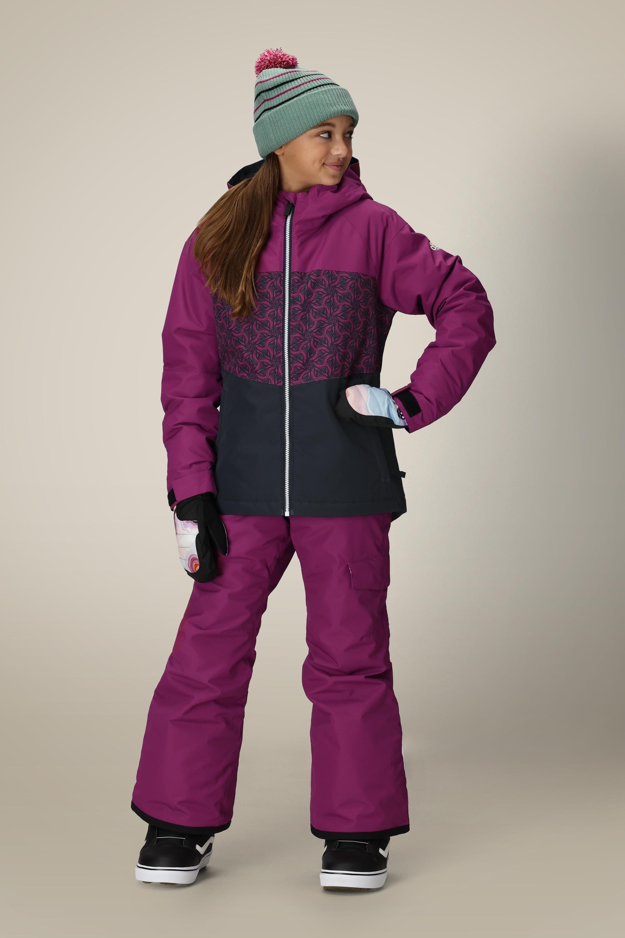 A young girl smiles, wearing the 686 Girls Athena Insulated Jacket in purple and black with matching snow pants, plus a green knit hat and gloves, looking off to the side against a plain background.