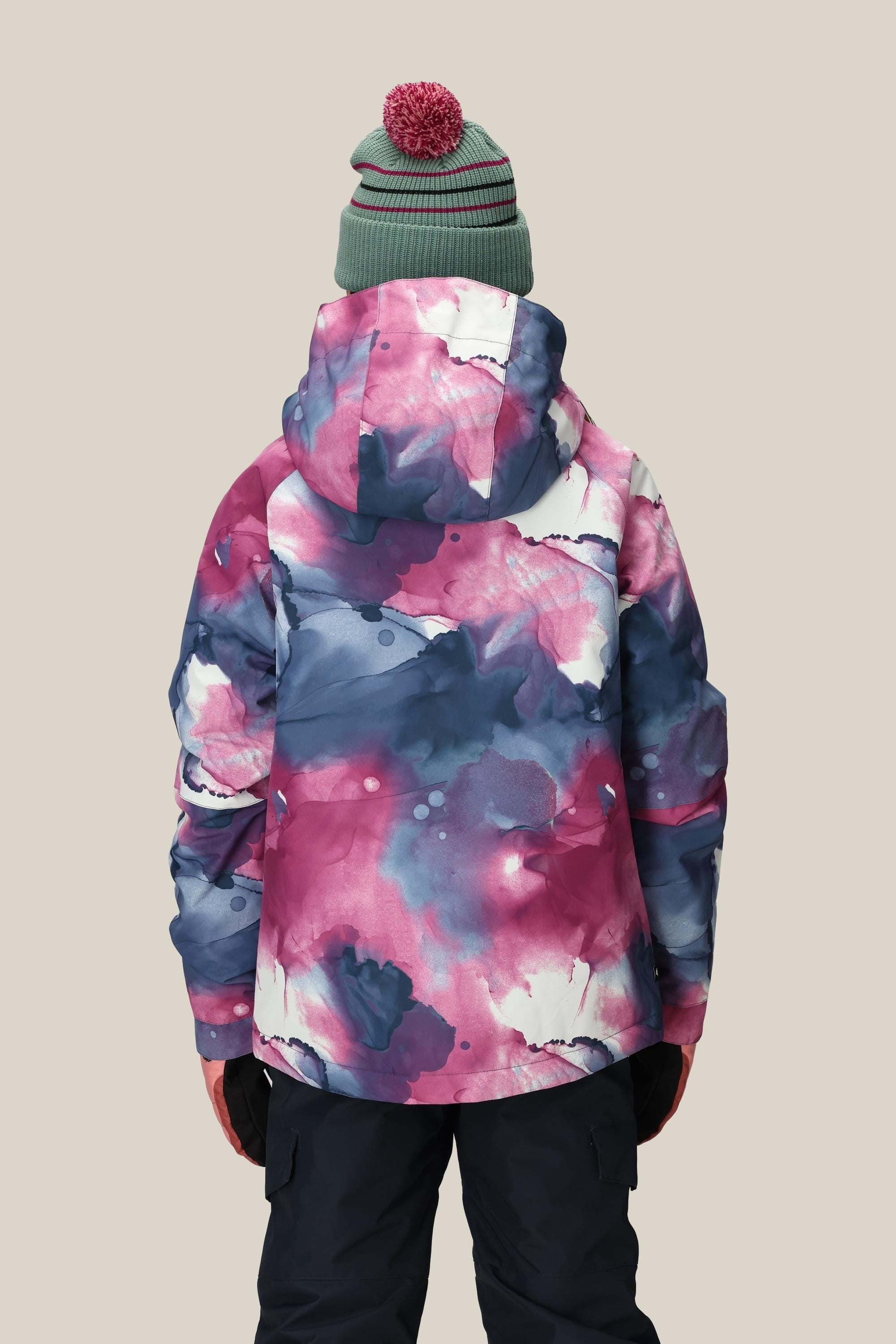 A person is shown from the back wearing the 686 Girls Hydra Insulated Jacket by 686 in bright pink, purple, and blue abstract patterns, paired with a green knit hat with pink stripes and pom-pom, and dark pants against a plain background.