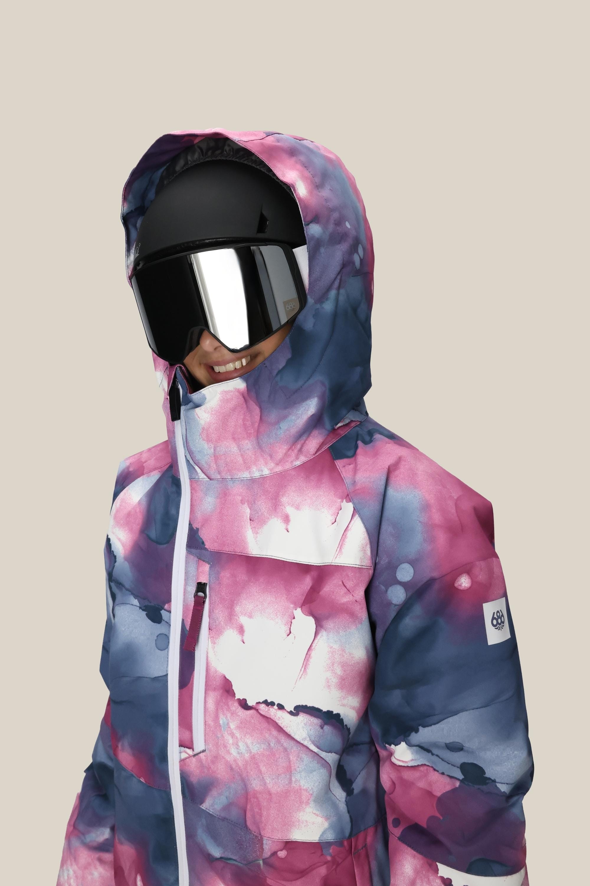 A girl in the 686 Girls Hydra Insulated Jacket with a vibrant watercolor pattern, black ski helmet, and reflective goggles smiles against a plain beige background.