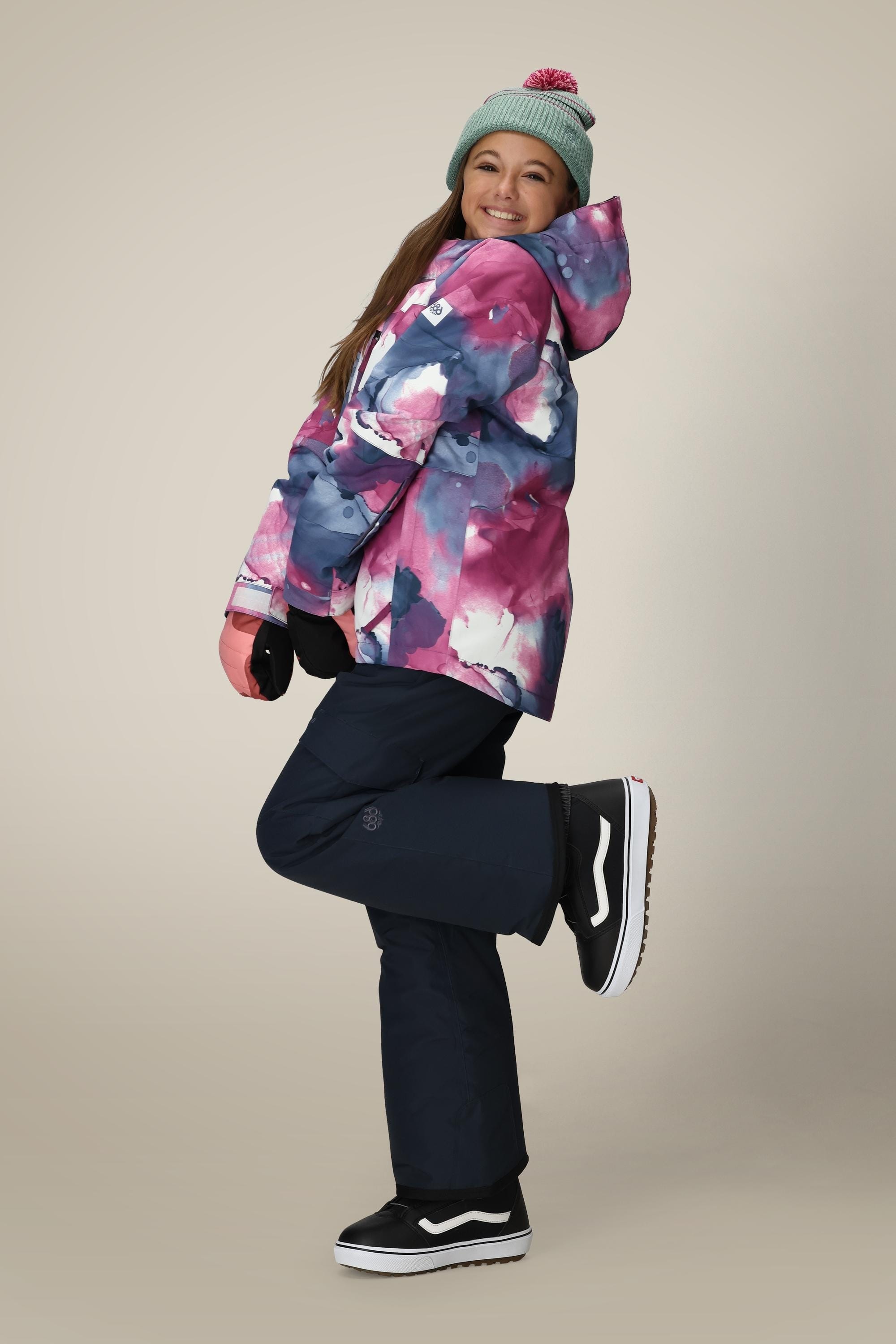 A smiling woman in a gray beanie, wearing the 686 Girls Hydra Insulated Jacket and black snow pants, poses playfully on one leg against a plain beige background.